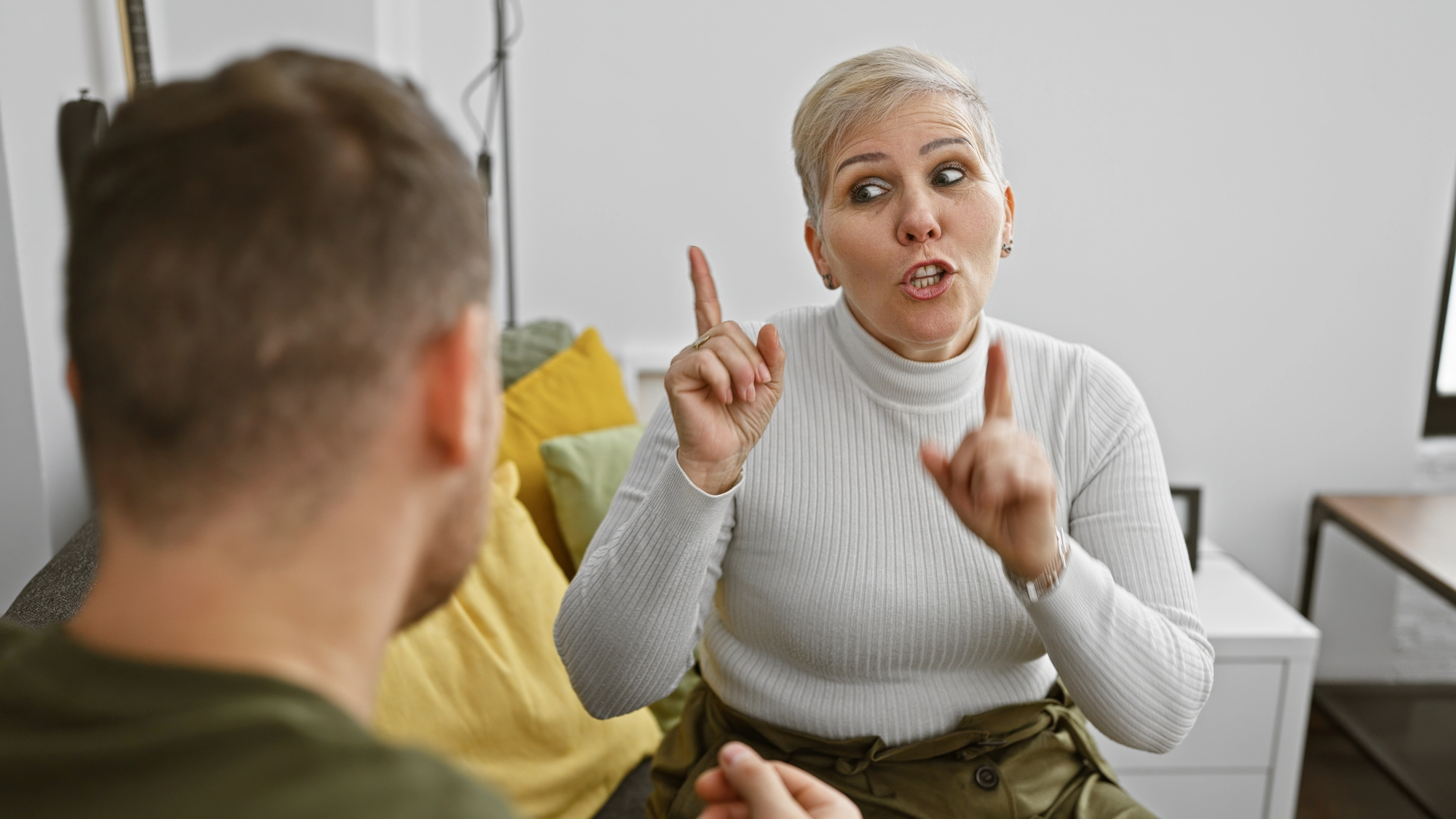 A woman with short hair and a white turtleneck gestures with both hands, pointing her index fingers upwards. She is engaged in a conversation with a man whose back is to the camera. They are indoors with pillows in the background.