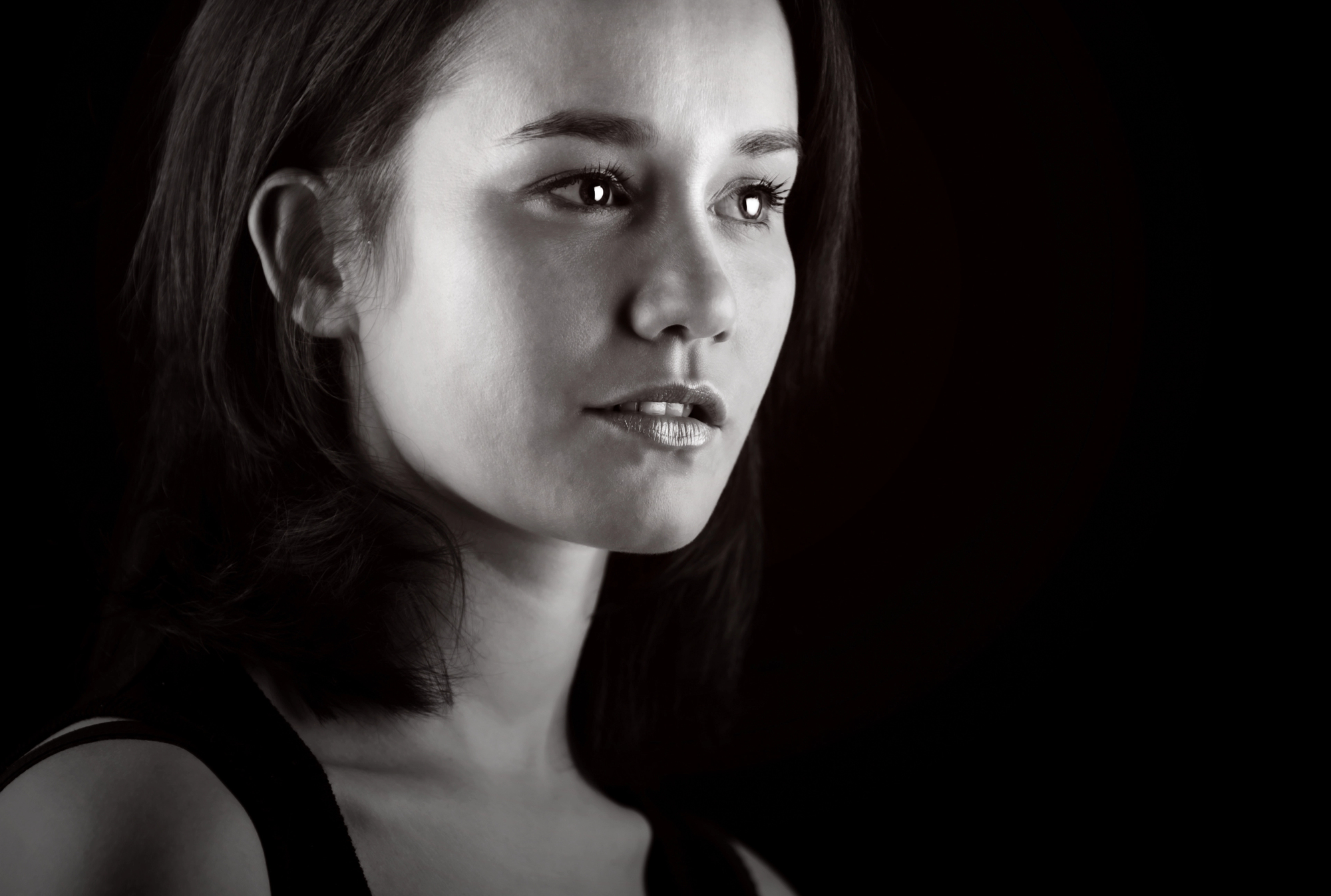 A black and white portrait of a person with shoulder-length hair, looking slightly to the side against a dark background. The lighting highlights their facial features, creating a contrast with the background.