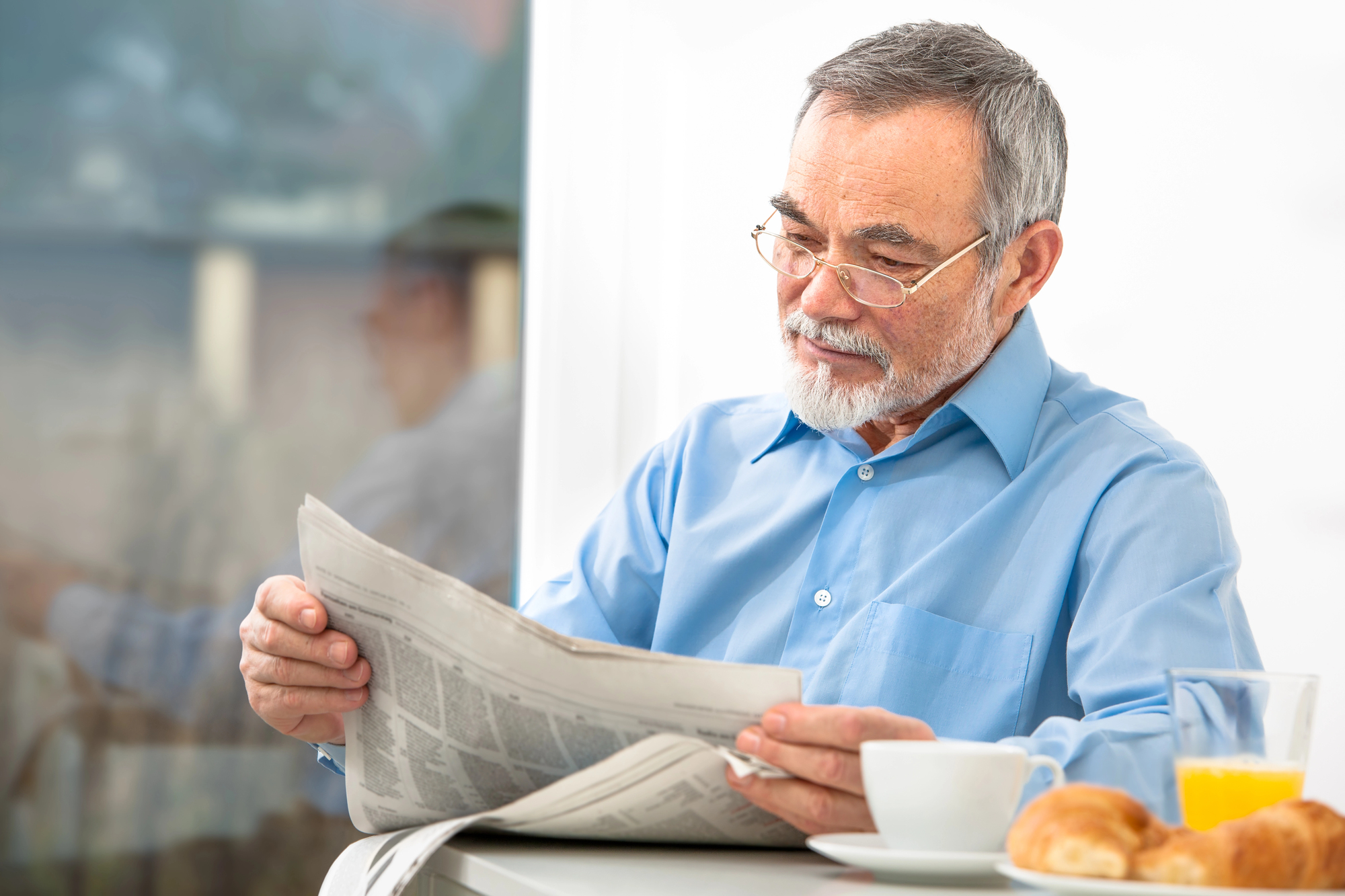 An older man with glasses and a beard is sitting at a table, reading a newspaper. A cup of coffee, a glass of orange juice, and some pastries are on the table. There is a window next to him reflecting his image.