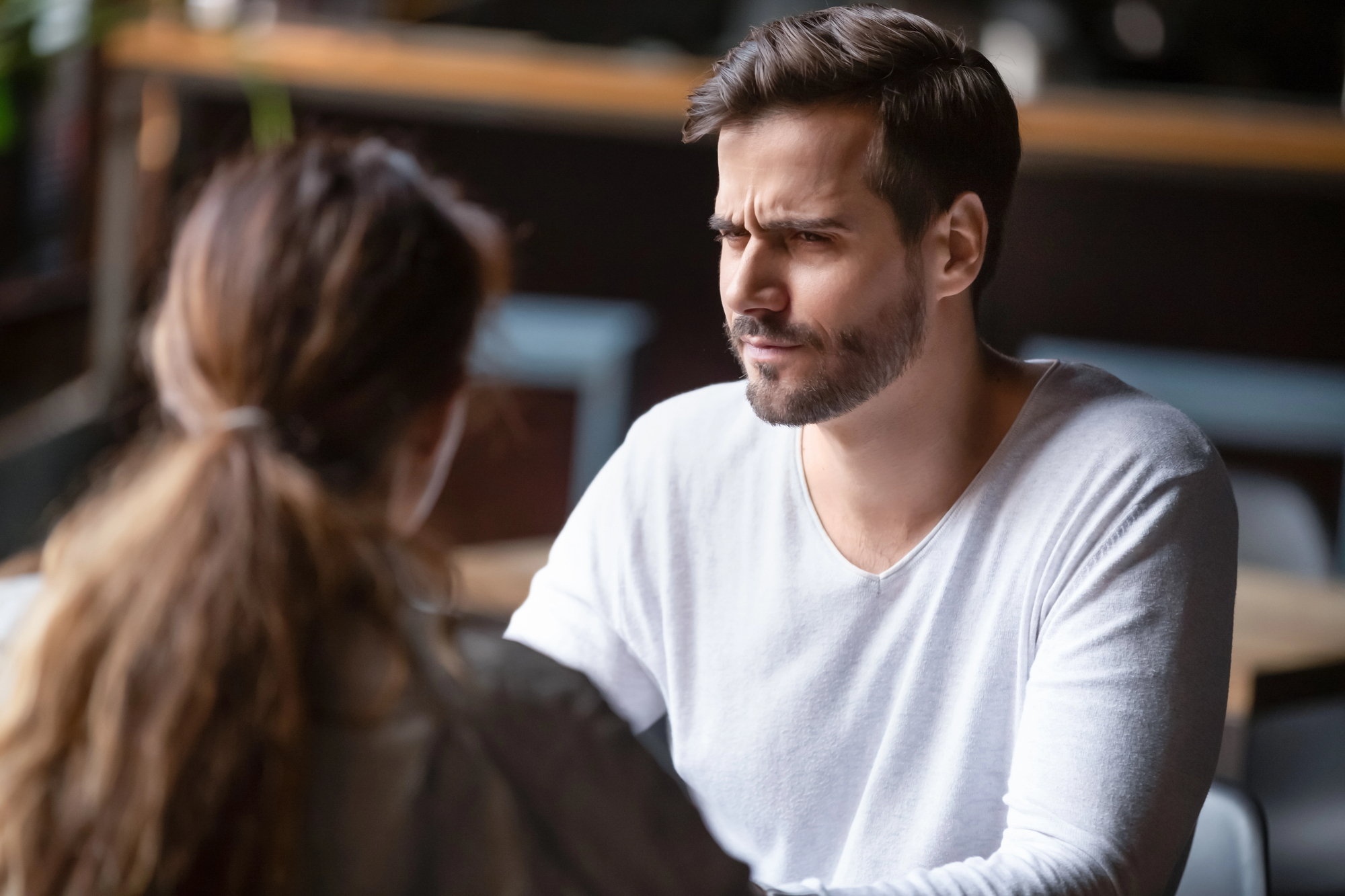 A man with a beard and wearing a white shirt sits at a table in conversation with a woman whose back is to the camera. His expression is thoughtful and slightly furrowed, as if engaged in a serious discussion. The setting appears to be a casual cafe.