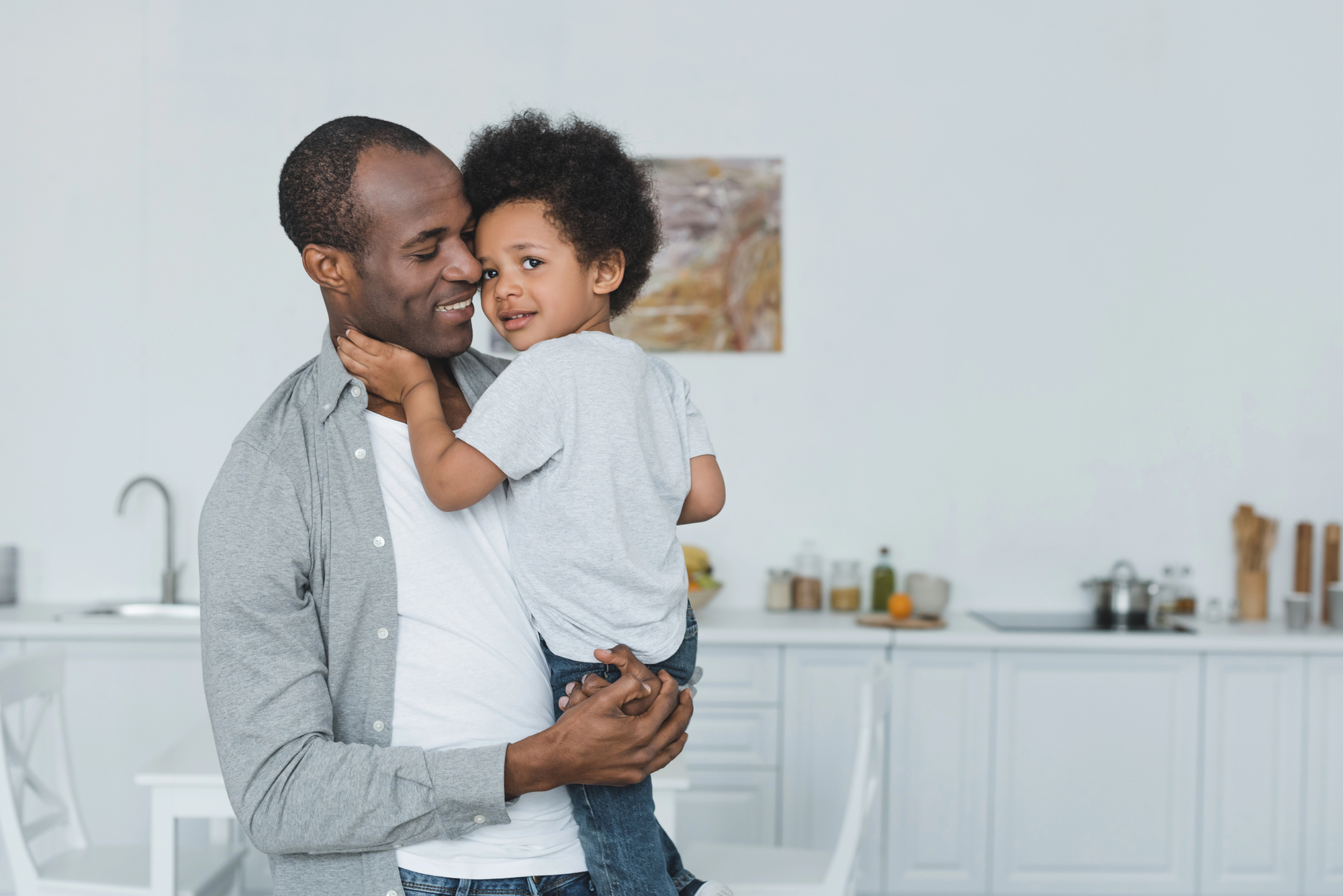 A smiling man holds a young child in his arms in a modern kitchen. Both are wearing casual grey shirts, and the child gently touches the man's cheek. The background shows a blurred countertop with kitchen items.