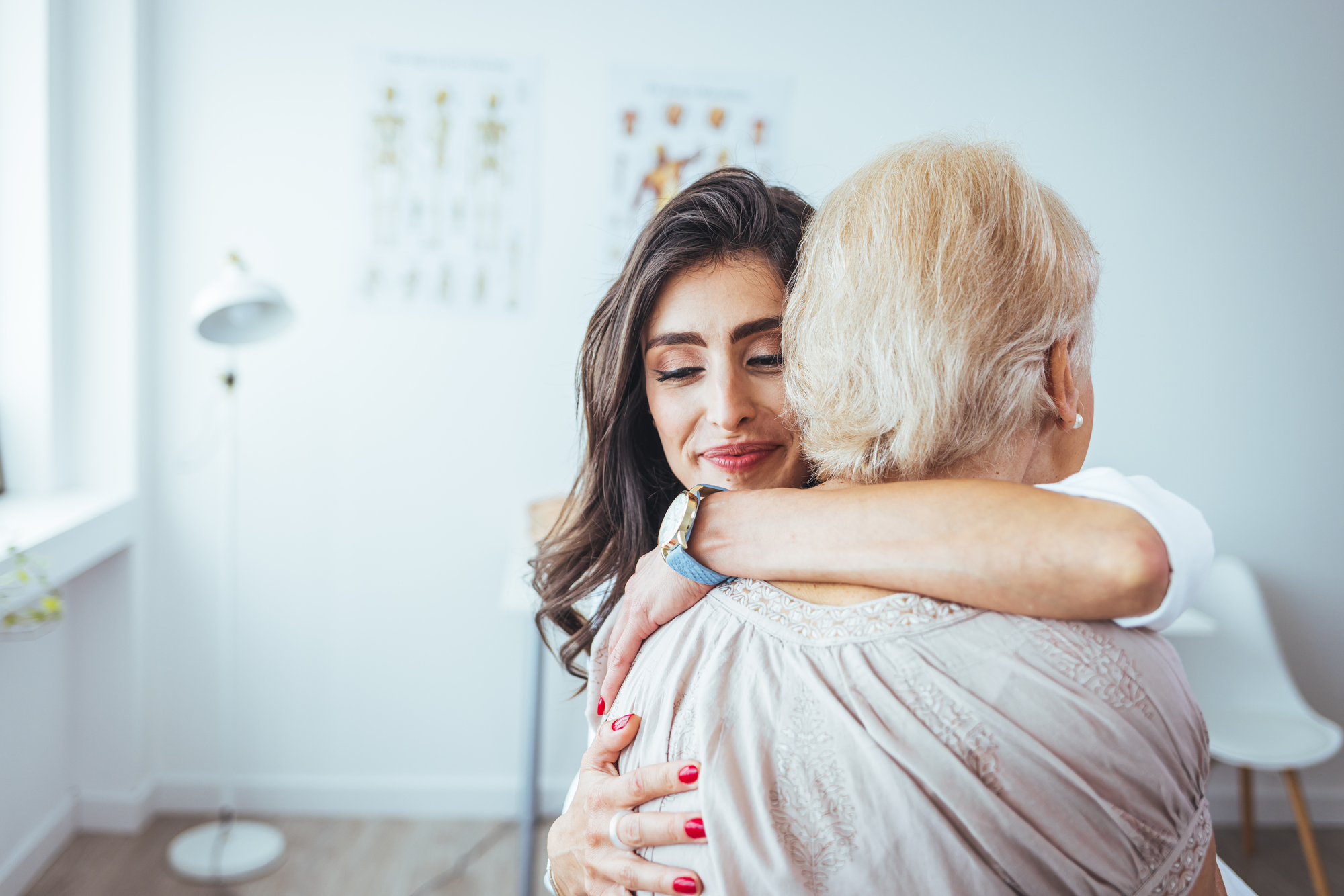 A young woman with long dark hair embraces an older woman with short blonde hair in a bright, white room. Both appear content and are sharing a close, warm hug. Charts are visible on the wall in the background.