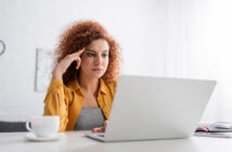 A woman with curly hair sits at a table, focused on her laptop. She wears a mustard yellow shirt and rests her head on her hand. A cup and an open notebook are on the table, and the background is bright and minimalistic.