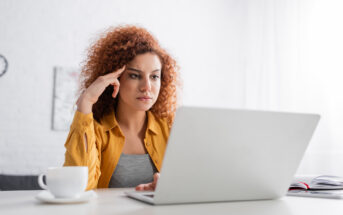 A woman with curly hair sits at a table, focused on her laptop. She wears a mustard yellow shirt and rests her head on her hand. A cup and an open notebook are on the table, and the background is bright and minimalistic.