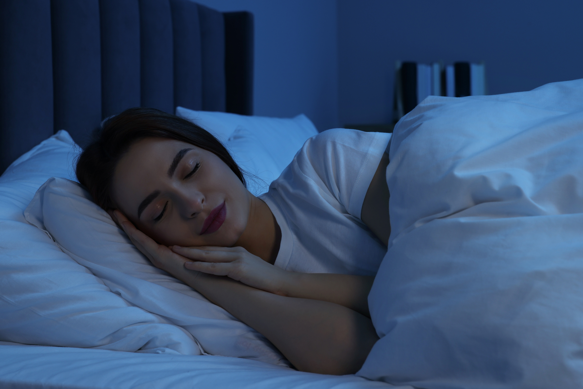 A person peacefully sleeping in bed, wrapped in a white blanket. The room is dimly lit, giving a sense of calm and tranquility. A few books are visible on a shelf in the background.