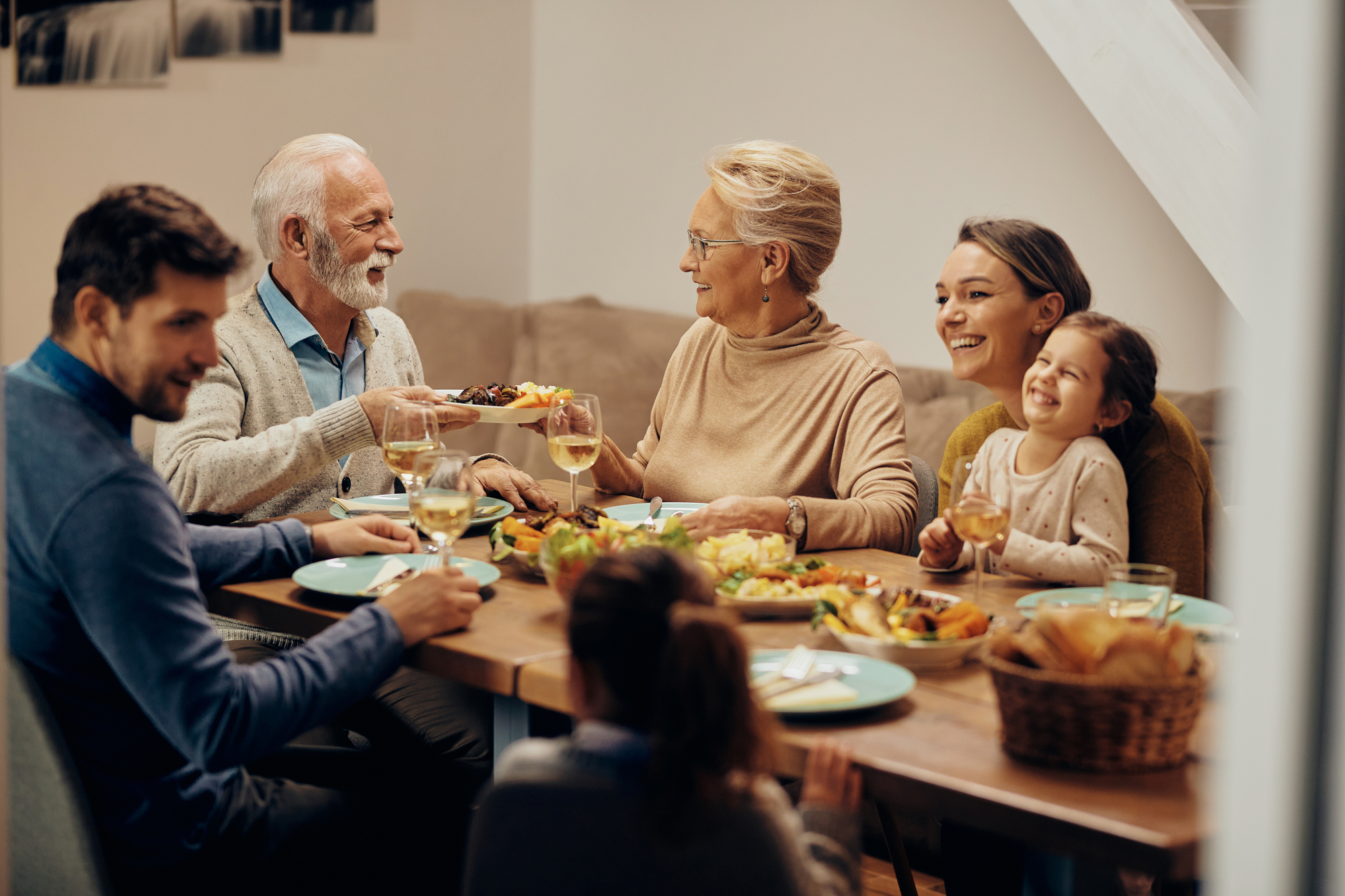 A group of five people, including an elderly couple, two adults, and a child, sit around a dining table sharing a meal. They are smiling and enjoying each other's company, with plates of food and glasses of wine in front of them.