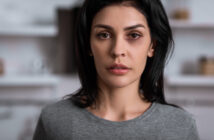 A woman with long dark hair and a serious expression looks directly at the camera. She is wearing a gray top, and the background appears to be a softly blurred indoor setting.