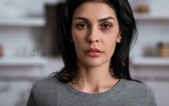 A woman with long dark hair and a serious expression looks directly at the camera. She is wearing a gray top, and the background appears to be a softly blurred indoor setting.