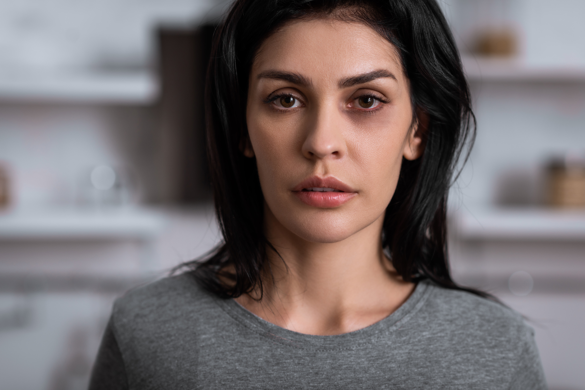 A woman with long dark hair and a serious expression looks directly at the camera. She is wearing a gray top, and the background appears to be a softly blurred indoor setting.