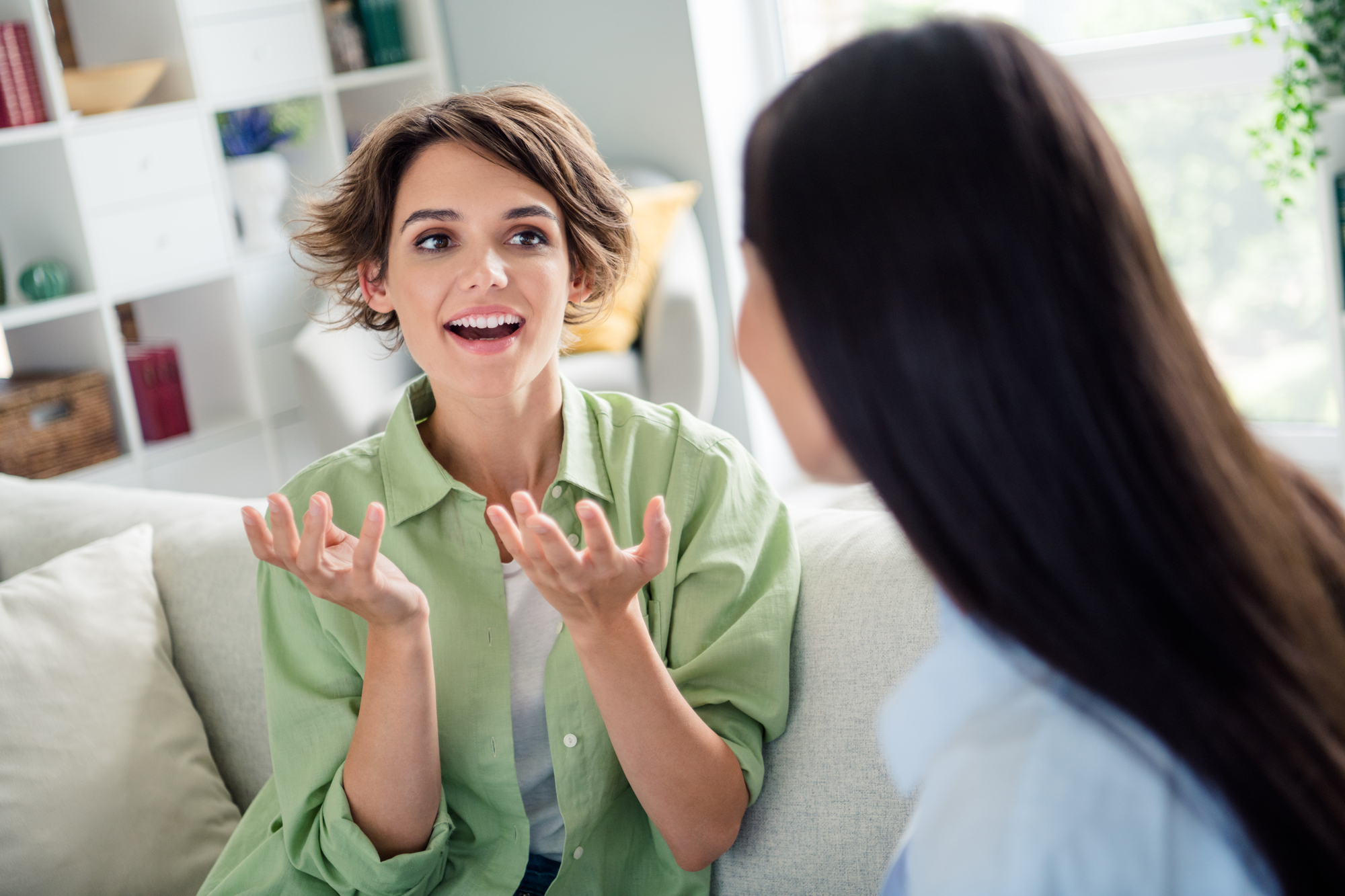 A woman with short hair wearing a green shirt is sitting on a couch, gesturing animatedly while talking to another woman with long dark hair. They are in a bright room with shelves and plants in the background.