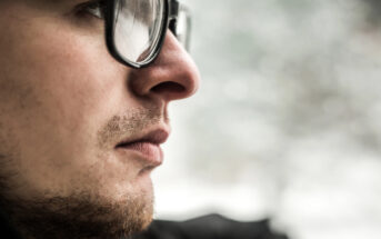 A close-up side profile of a person wearing glasses, with a focused expression. The background is blurred, suggesting an outdoor, possibly snowy setting. The image emphasizes facial details and glasses.