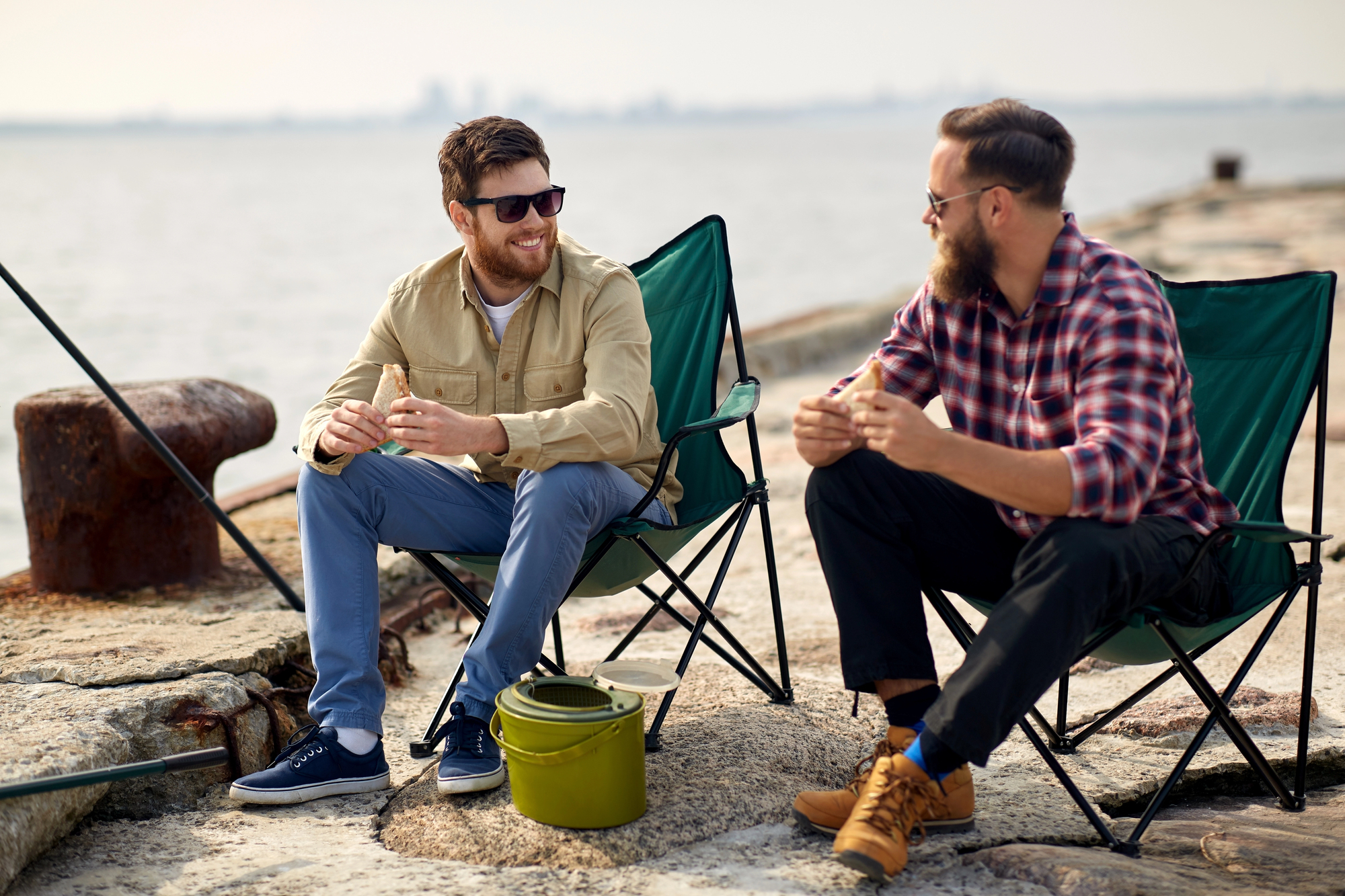 Two men are seated in camping chairs by a waterfront. They are smiling, wearing casual clothing, and appear to be engaged in conversation. Fishing poles are positioned nearby, and a green bucket sits on the ground between them. The background features a calm body of water.