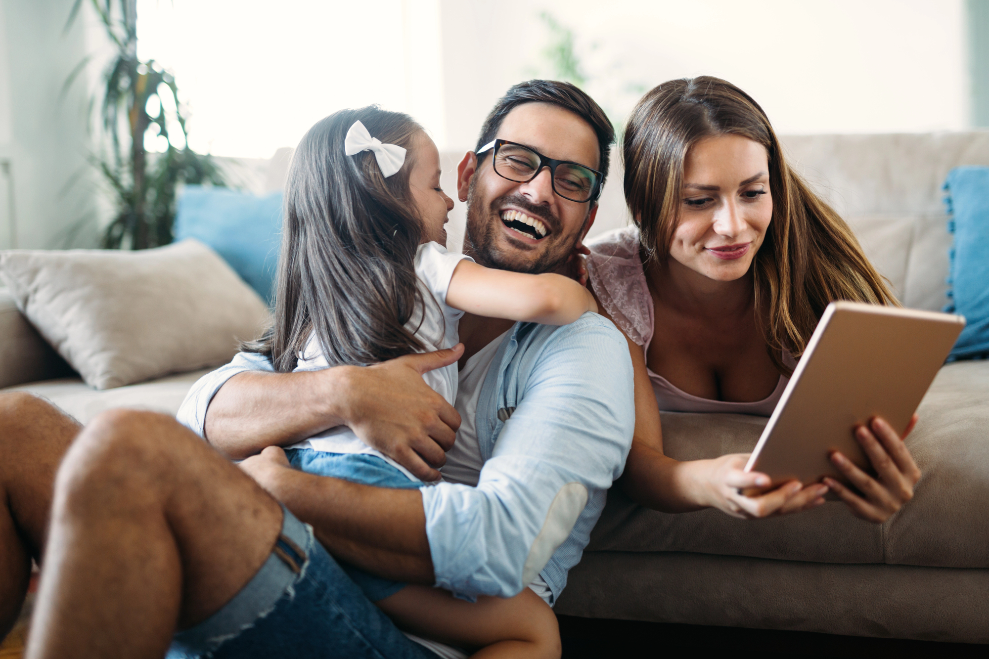 A happy family is spending time together at home. A man is sitting on the floor, laughing and hugging a young girl. A woman is lying next to them on the couch, smiling and holding a tablet. The room has a cozy and relaxed atmosphere.