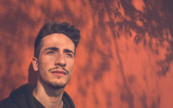 Young man with short hair and a mustache gazes into the distance against a sunlit orange wall with leaf shadows. The warm light creates a serene and thoughtful atmosphere.