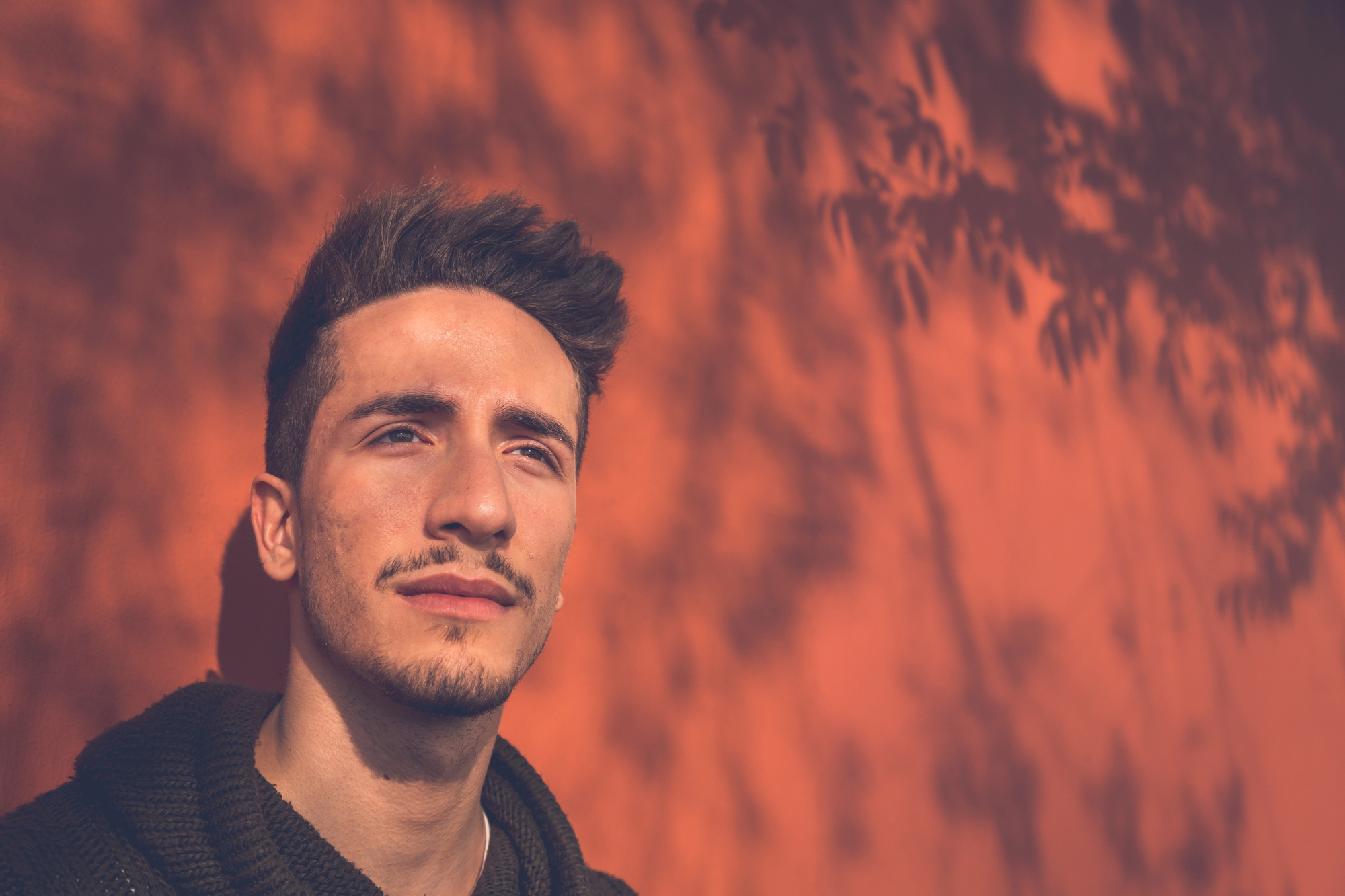 Young man with short hair and a mustache gazes into the distance against a sunlit orange wall with leaf shadows. The warm light creates a serene and thoughtful atmosphere.