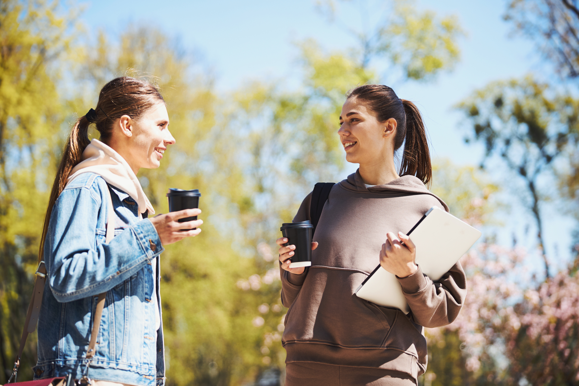 Two women stand outdoors on a sunny day, holding coffee cups and chatting. One is dressed in a denim jacket, the other in a brown hoodie while holding a tablet. Blossoming trees are in the background, indicating springtime.