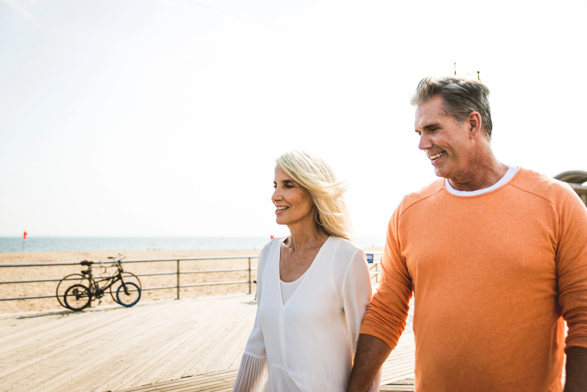 A couple walks hand in hand on a sunny boardwalk by the beach. The man is wearing an orange sweater, and the woman is wearing a white blouse. Two bicycles are parked by a railing in the background. The sky is clear and bright.