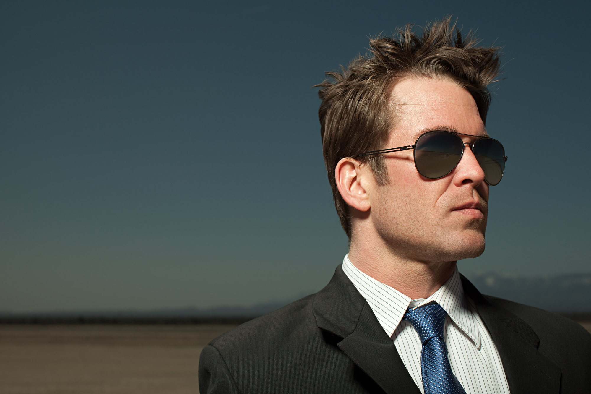 Man wearing sunglasses and a suit with a blue tie stands confidently against a clear blue sky backdrop.