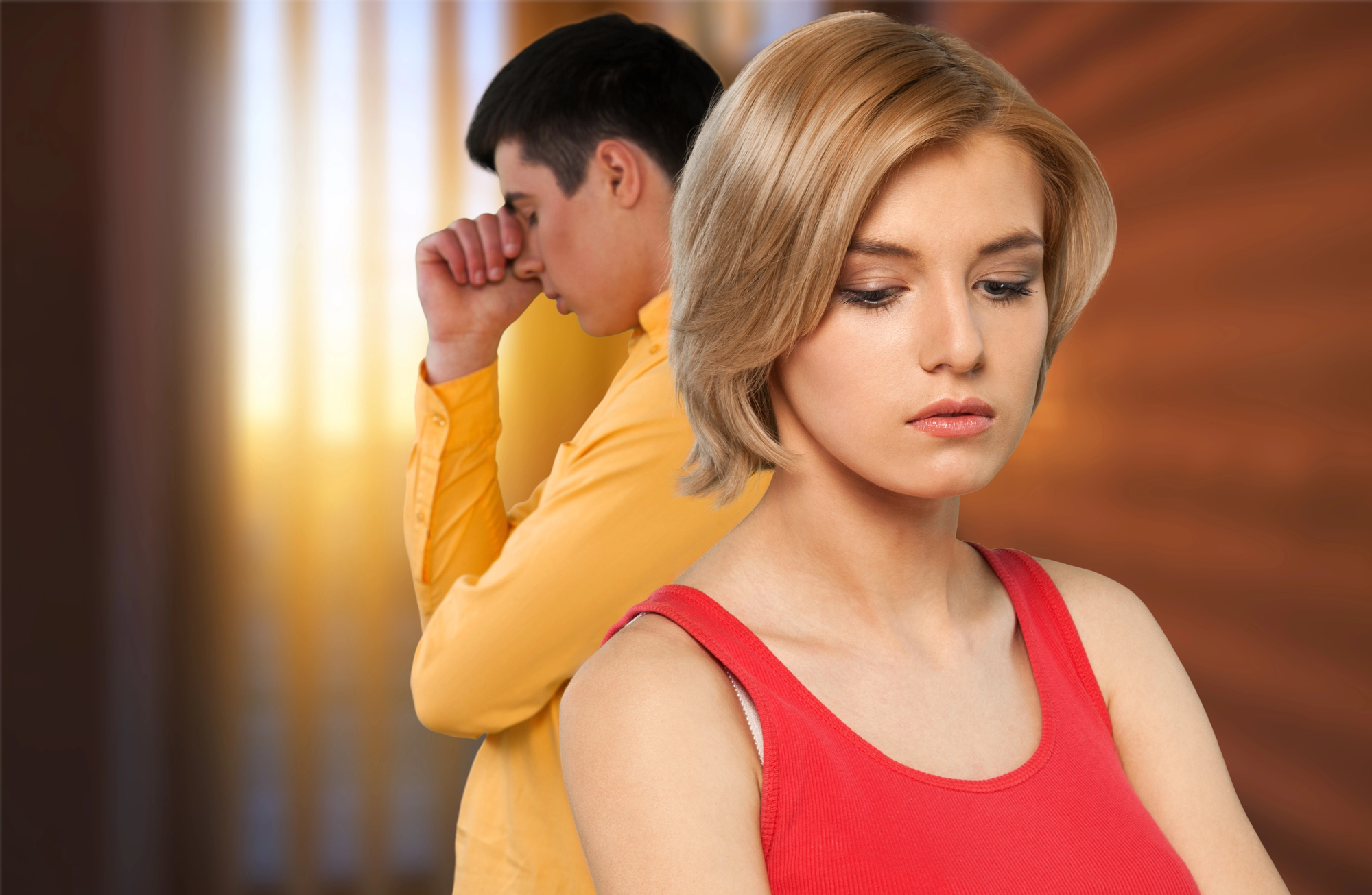 A woman with short blonde hair in a red tank top looks down, appearing thoughtful or upset. In the background, a man in a yellow shirt is turned away, resting his hand on his forehead. The scene suggests emotional tension.