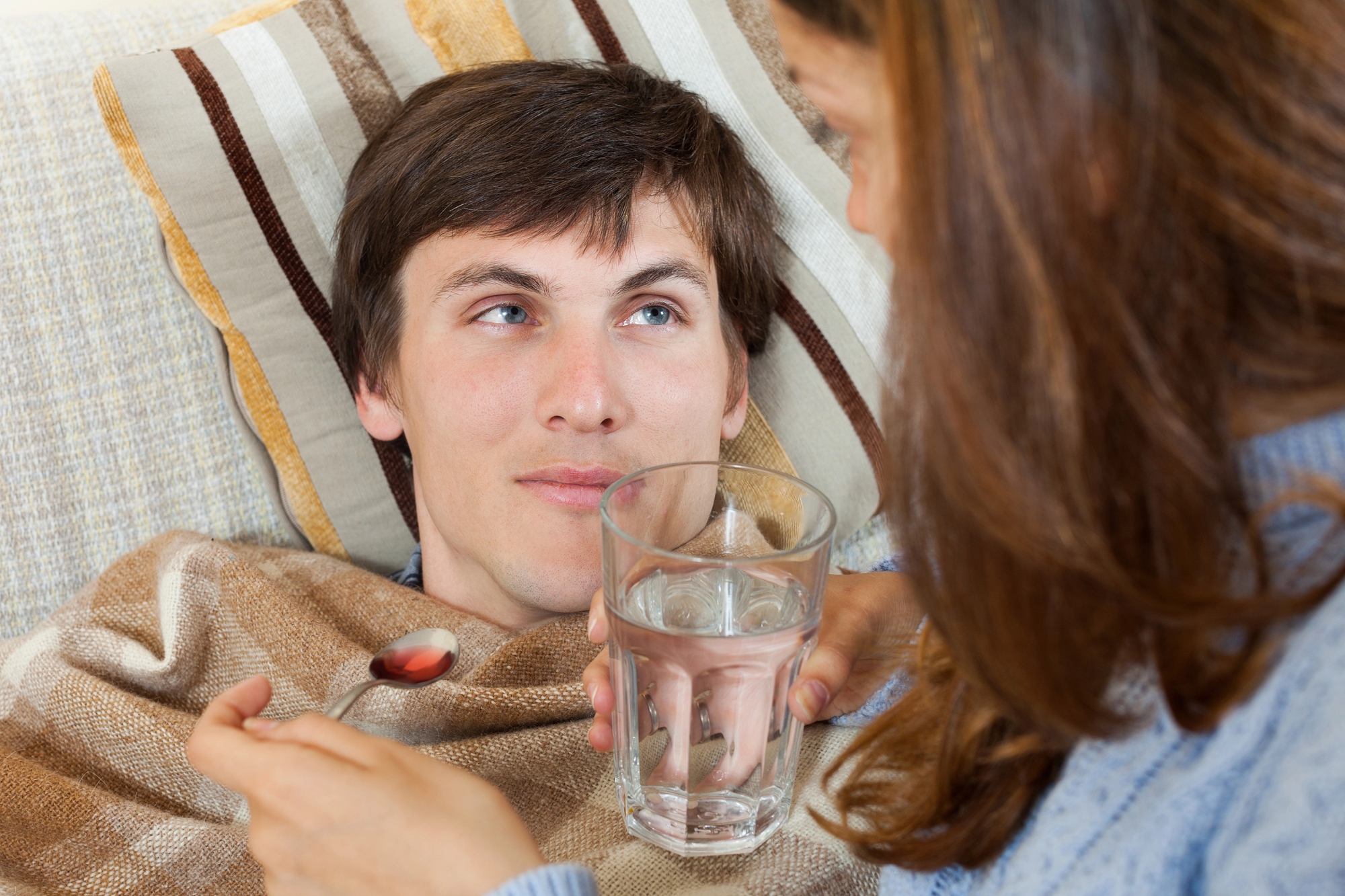 A young man lies on a couch, wrapped in a blanket, looking up at a woman. She is offering him a glass of water and a spoonful of medicine. The setting is cozy, suggesting care and attention.