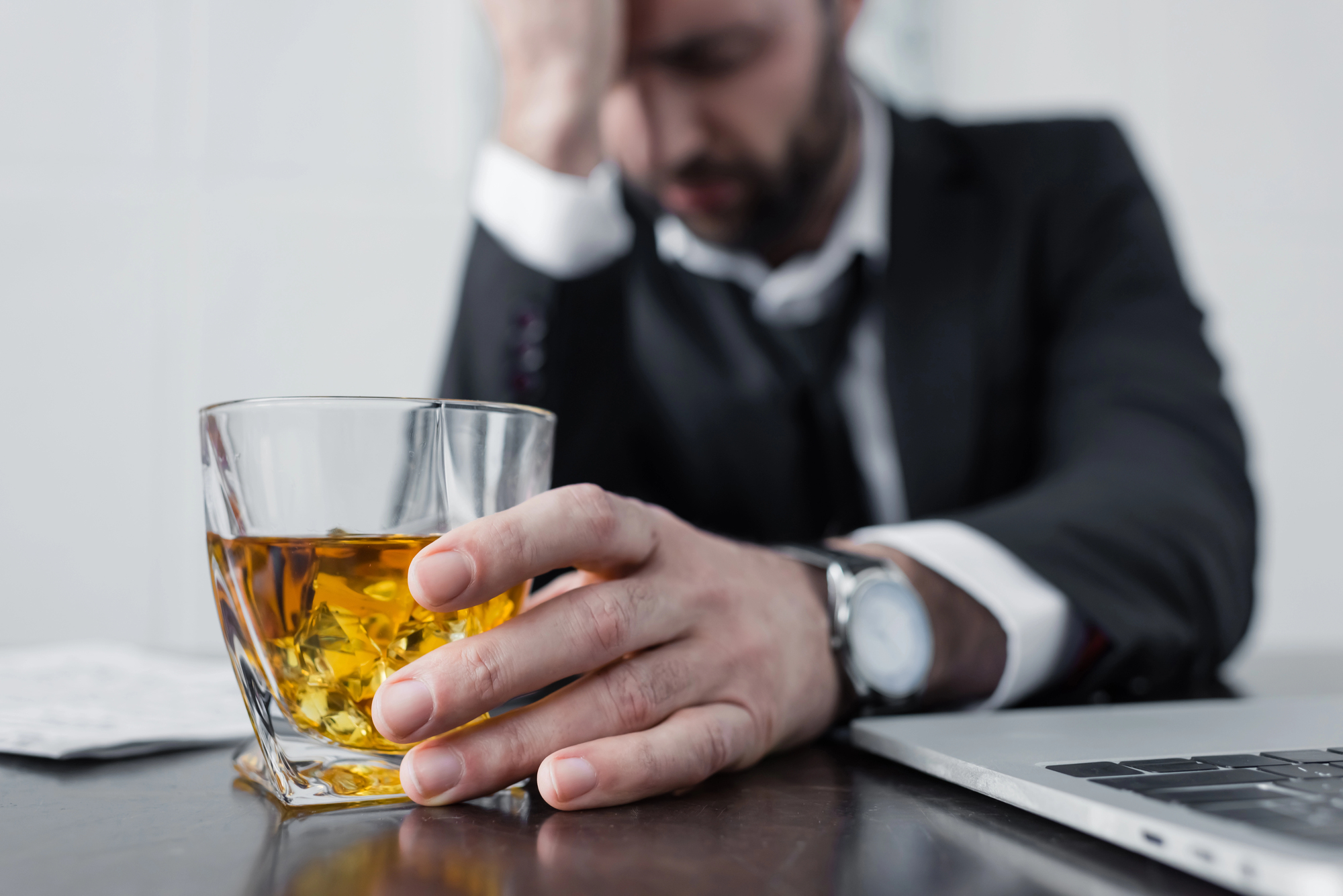 A person sitting at a desk, holding a glass of amber liquid, possibly whiskey. The person appears stressed, with hand on forehead. A laptop and papers are on the desk. The person is wearing a suit and watch.