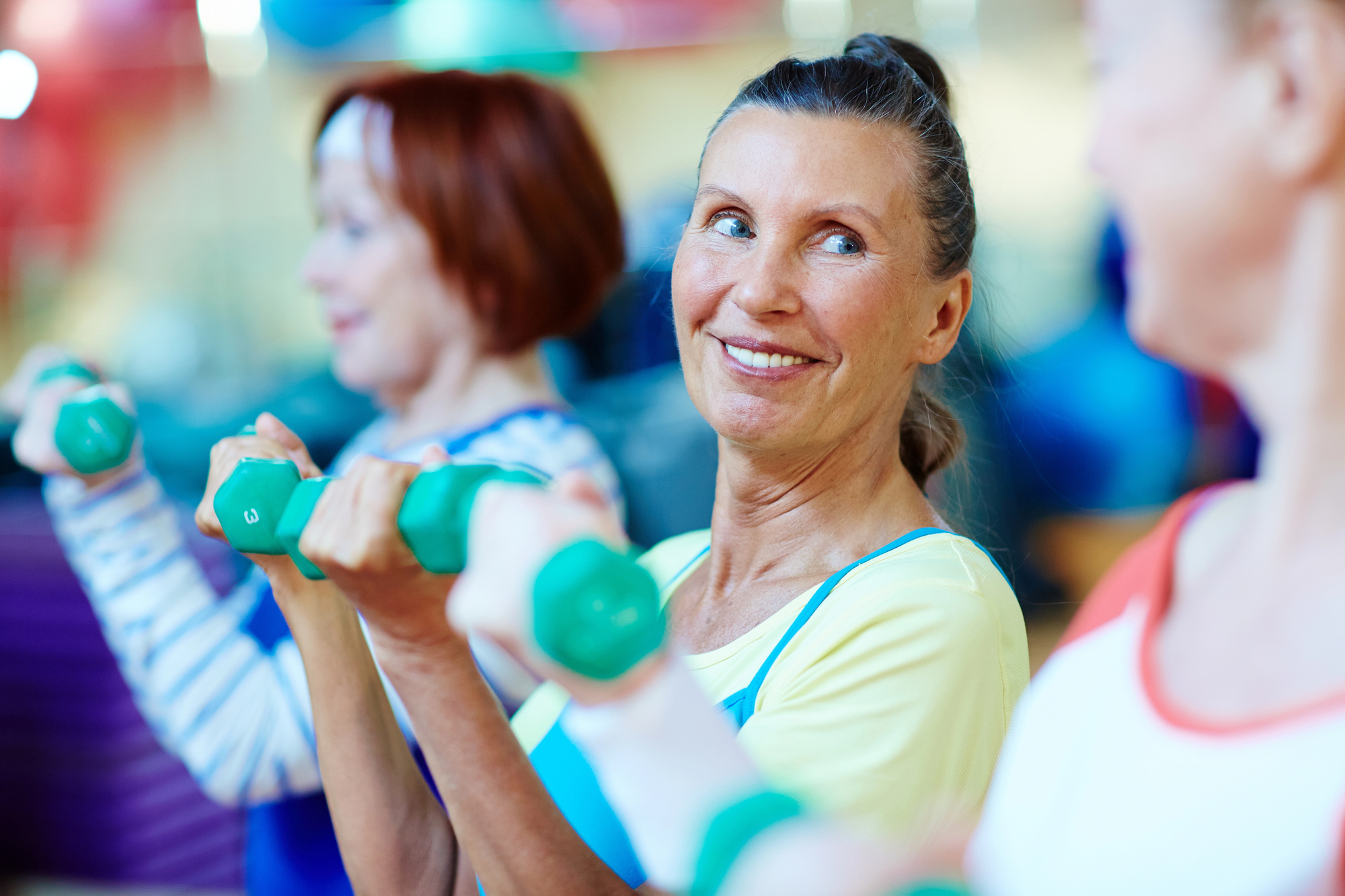 A group of women exercising in a gym, holding green dumbbells and smiling. The focus is on a woman in the center looking at someone beside her. The background is blurred, with two others visible in workout attire.