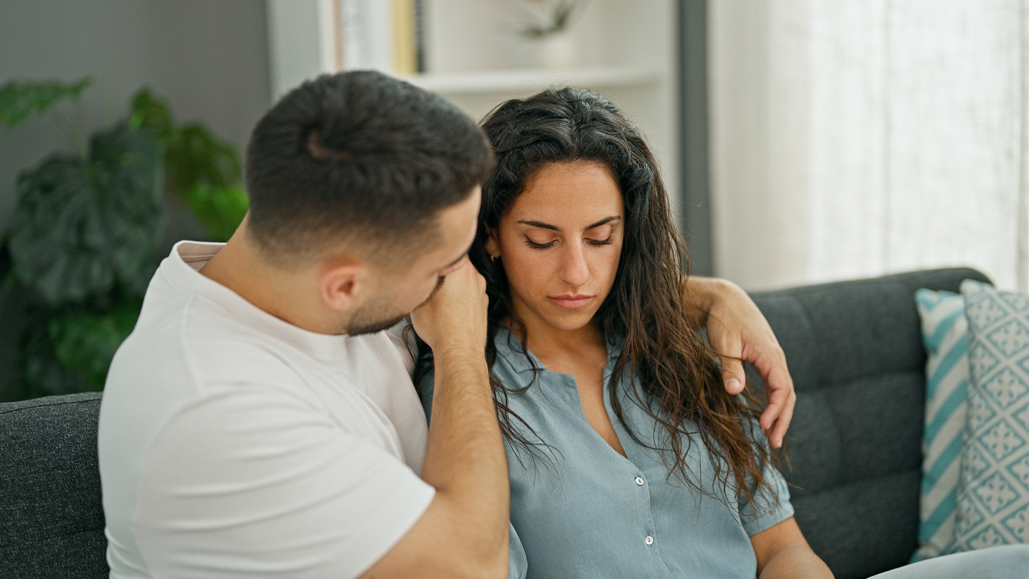 A man and a woman sit on a couch; the man has his arm around the woman's shoulder, offering comfort. The woman, with long dark hair, looks downward with a sad expression. The background shows plants and soft furnishings.