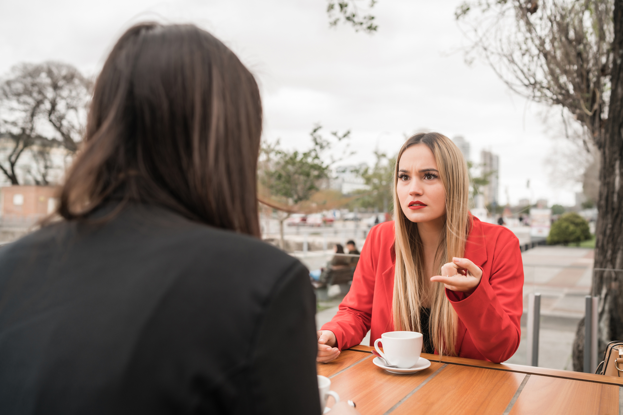 Two women having a serious conversation at an outdoor café. One woman, dressed in a red jacket, is gesturing while talking, and the other woman, with her back to the camera, listens. Each has a cup of coffee in front of them. Trees and buildings are in the background.