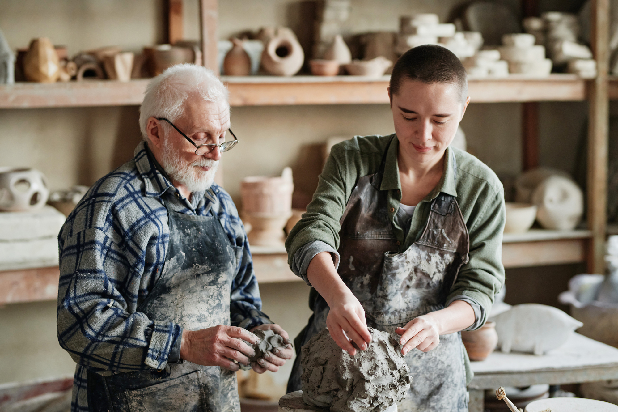 An older man and a younger person work together on pottery in a studio, shaping clay and wearing aprons. Shelves with pottery pieces are visible in the background.