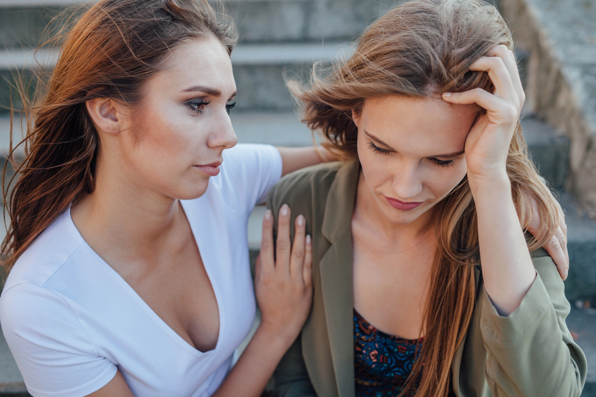 Two young women sitting on steps. One appears upset, with her head in her hand, while the other gently supports her with a hand on her shoulder. Their expressions convey concern and empathy in an outdoor setting.