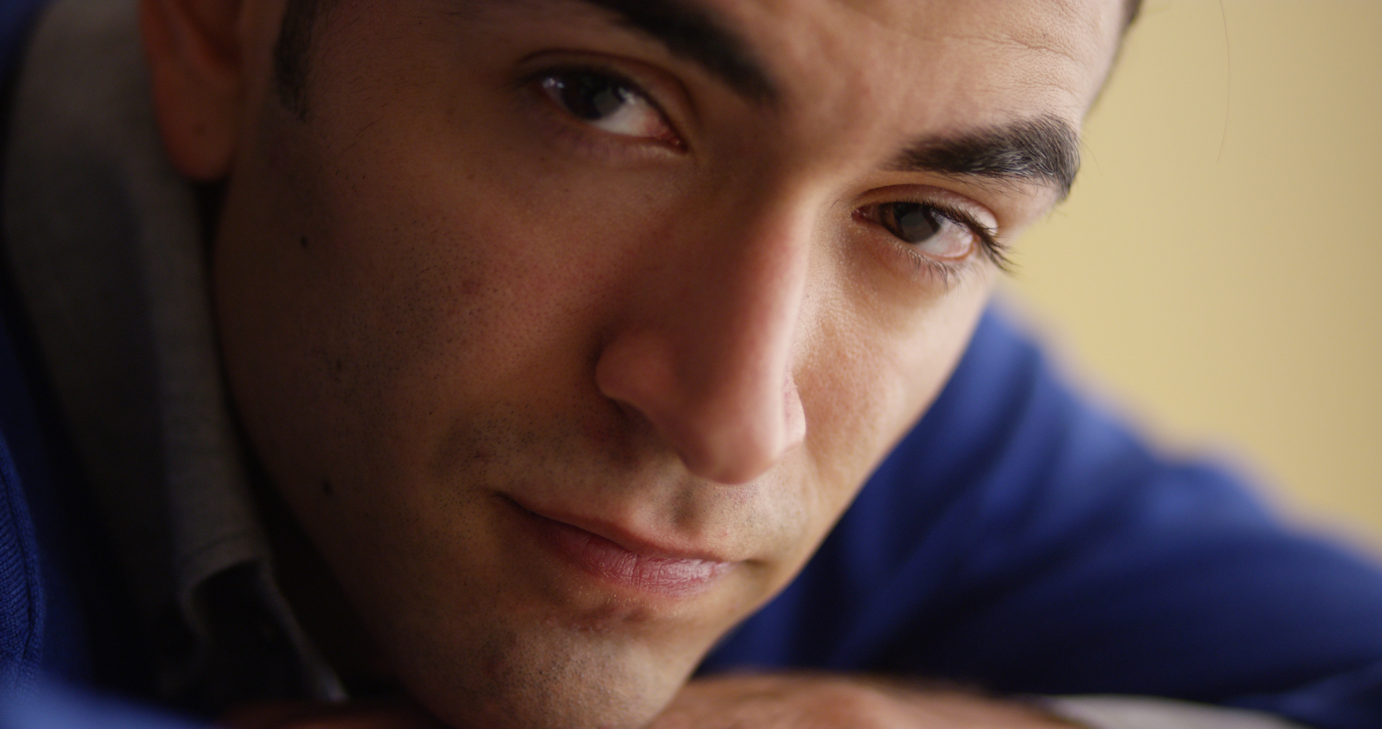 Close-up of a man with short dark hair and a blue sweater, looking directly at the camera. He has a calm expression with a slight smile, resting his chin on his hand against a soft, out-of-focus background.