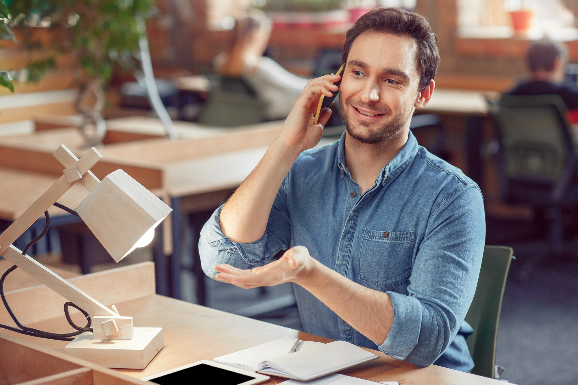 A man in a denim shirt is sitting at a desk in a modern office, talking on the phone and gesturing with his hand. A notebook and a tablet are in front of him, and a wooden desk lamp is on the side. Other people work in the background.