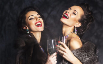 Two women, dressed in elegant fur coats and sparkling jewelry, are laughing and raising glasses of champagne. Their hairstyles and makeup are glamorous, and they are set against a dark, textured background.