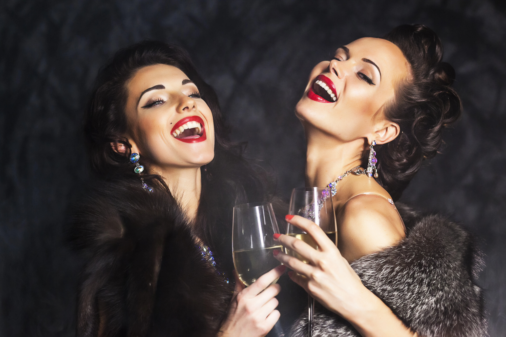 Two women, dressed in elegant fur coats and sparkling jewelry, are laughing and raising glasses of champagne. Their hairstyles and makeup are glamorous, and they are set against a dark, textured background.