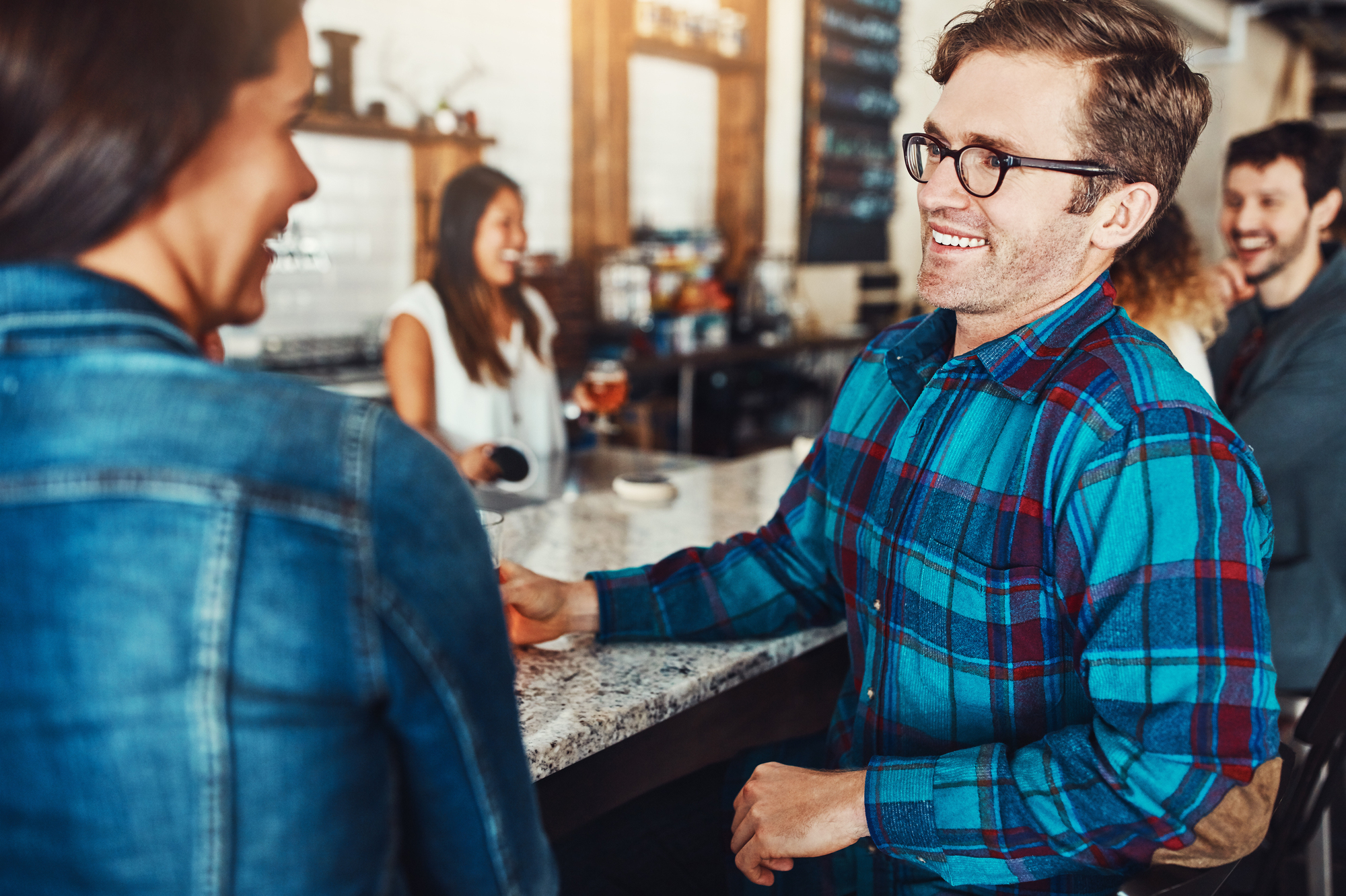 A man wearing glasses and a blue plaid shirt is sitting and smiling at a woman in a denim jacket at a bar counter, while two other people in the background are also smiling and talking. The setting is casual and relaxed with a warm ambiance.