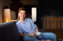 A woman in a blue striped shirt and jeans sits comfortably on a sofa, holding a mug and smiling. The background features a warmly lit interior with wooden accents and bar stools.