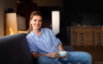 A woman in a blue striped shirt and jeans sits comfortably on a sofa, holding a mug and smiling. The background features a warmly lit interior with wooden accents and bar stools.