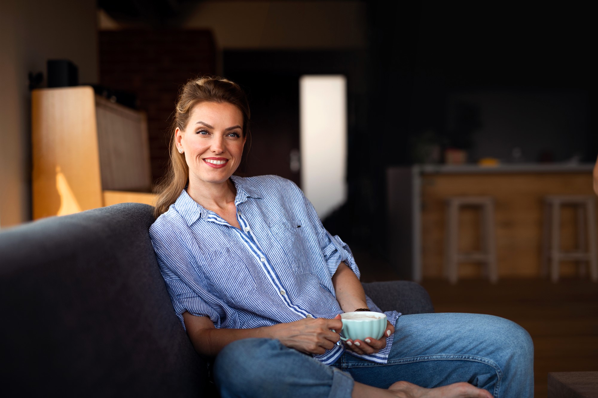 A woman in a blue striped shirt and jeans sits comfortably on a sofa, holding a mug and smiling. The background features a warmly lit interior with wooden accents and bar stools.