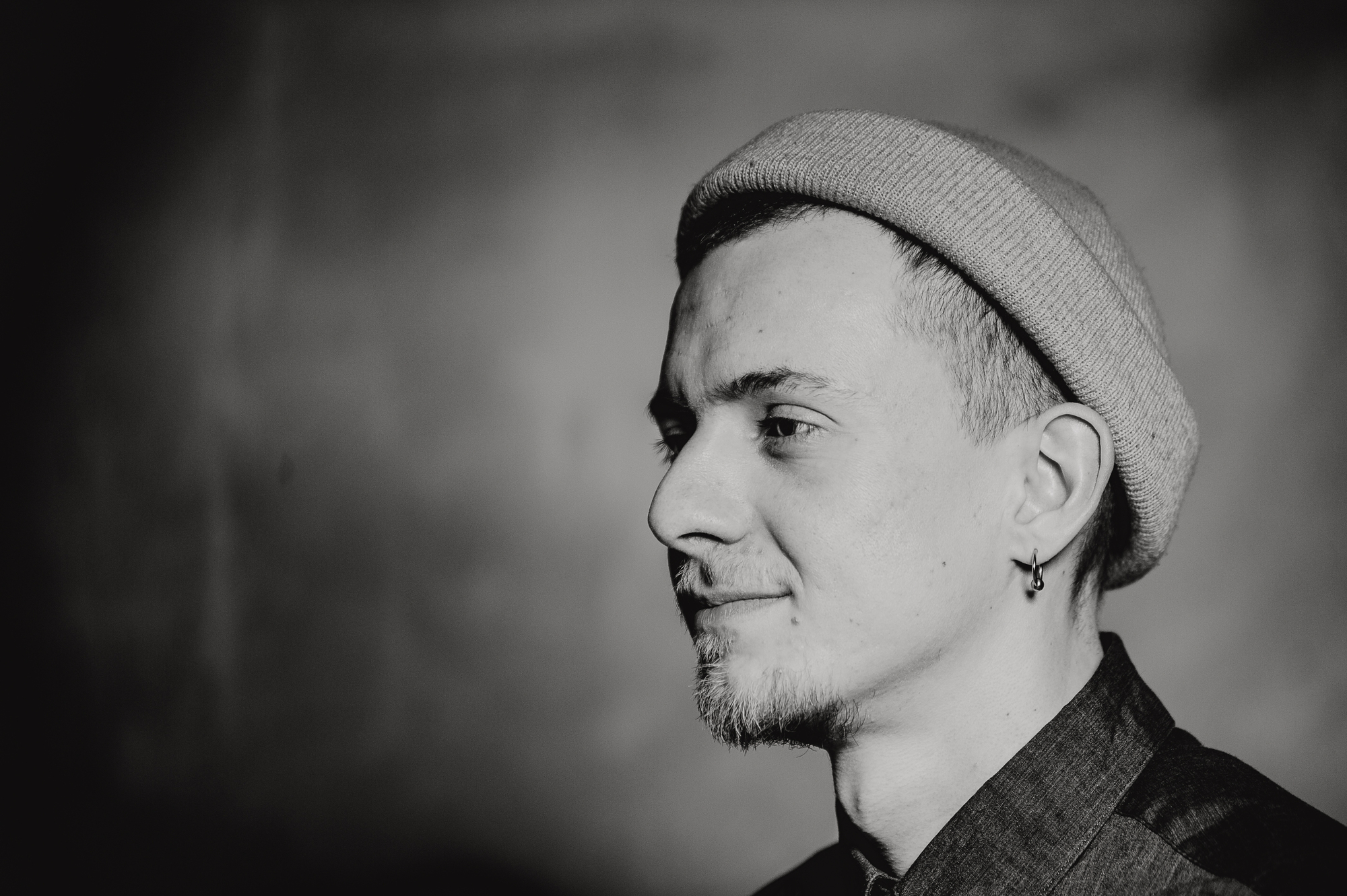 A black-and-white portrait of a person with a light beard and mustache, wearing a beanie and an earring. The person is looking to the right, with a slight smile. The background is soft-focused and neutral, emphasizing the person’s profile.