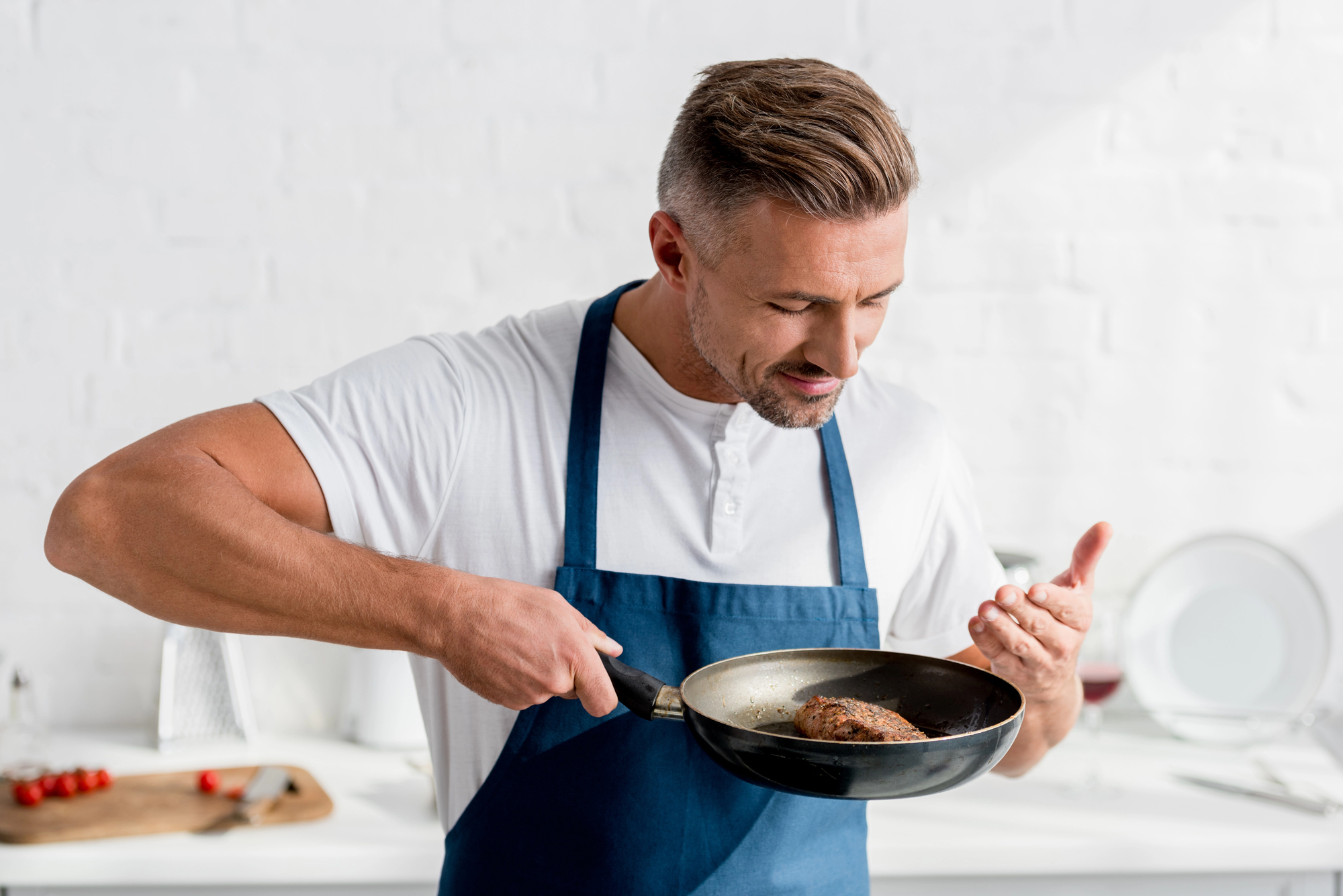 A man in a white shirt and blue apron is cooking in a bright kitchen. He holds a frying pan with a piece of meat, inspecting it closely with a focused expression. Plates and kitchen items are blurred in the background.