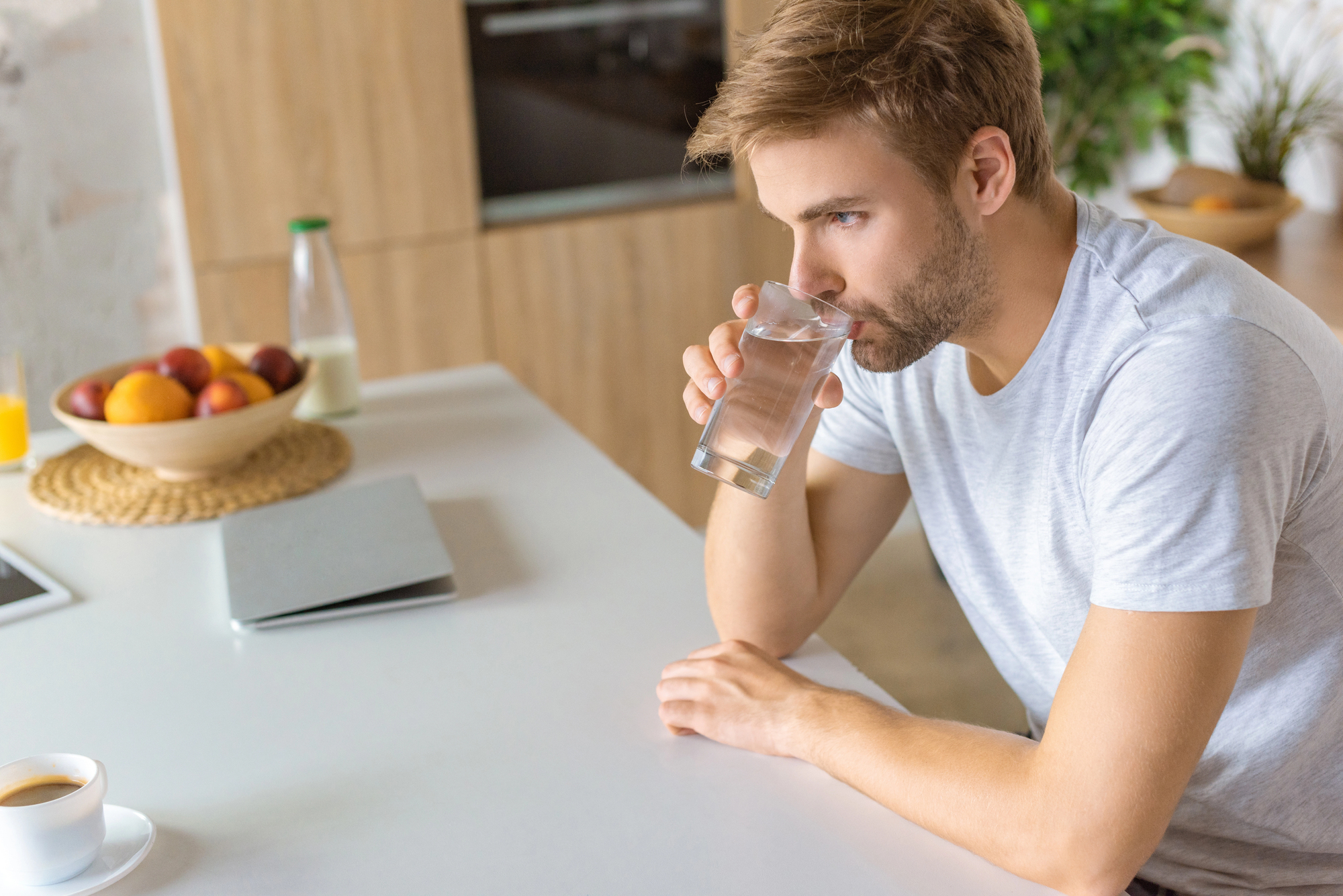 A man in a light gray t-shirt sits at a kitchen counter, drinking a glass of water. On the counter, there's a laptop, a cup of coffee, and a fruit bowl with oranges and apples. The background has kitchen cabinets and a green plant.