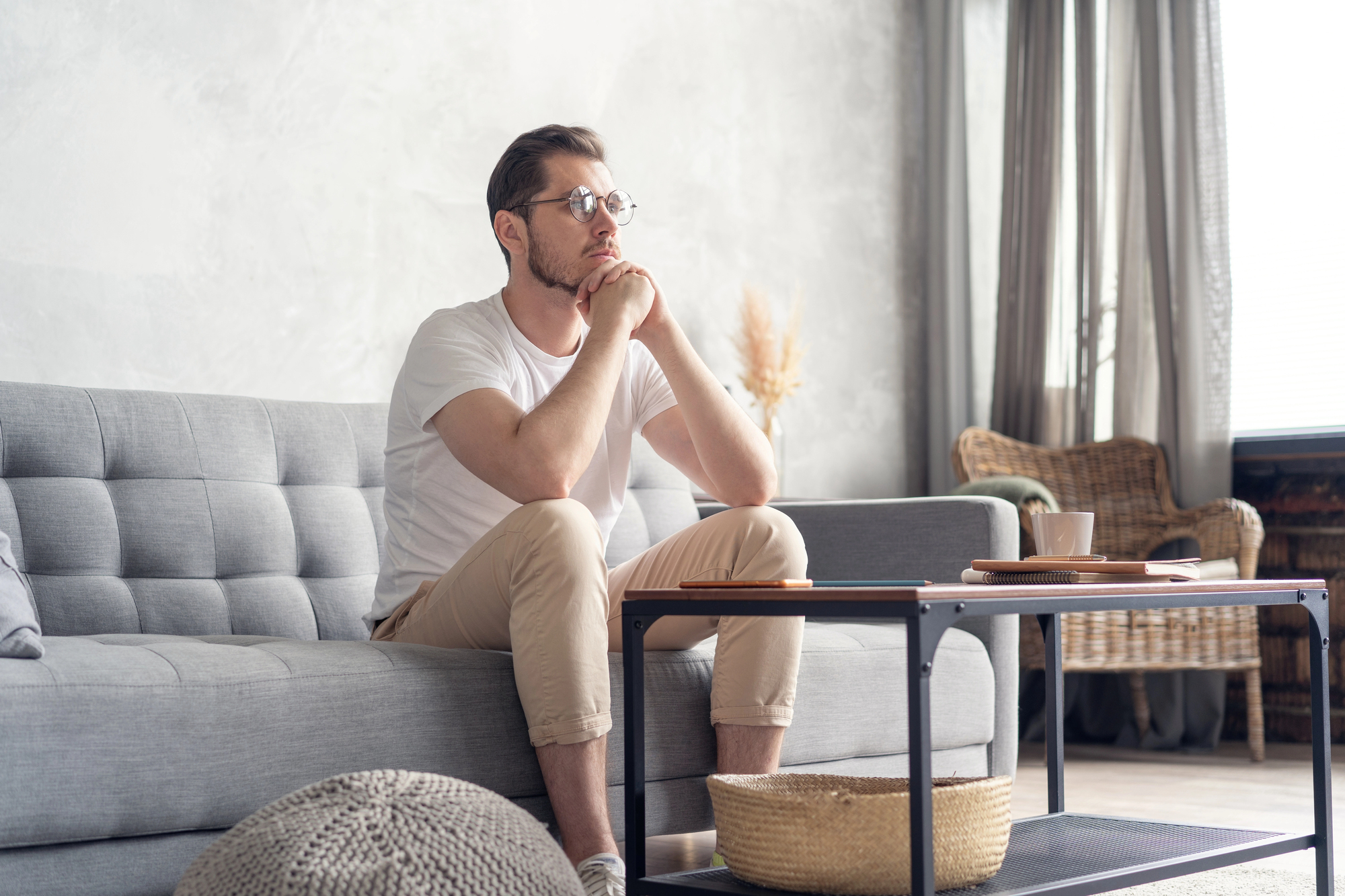 A man with glasses and a beard sits thoughtfully on a gray couch in a living room. He is resting his chin on his hands, elbows on his knees. The room has a modern decor with a coffee table, wicker chair, potted plants, and large windows letting in natural light.