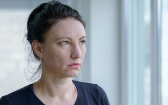 A woman with dark hair, tied back, stands by a window wearing a black top. She looks thoughtfully out the window, with soft natural light illuminating her face.