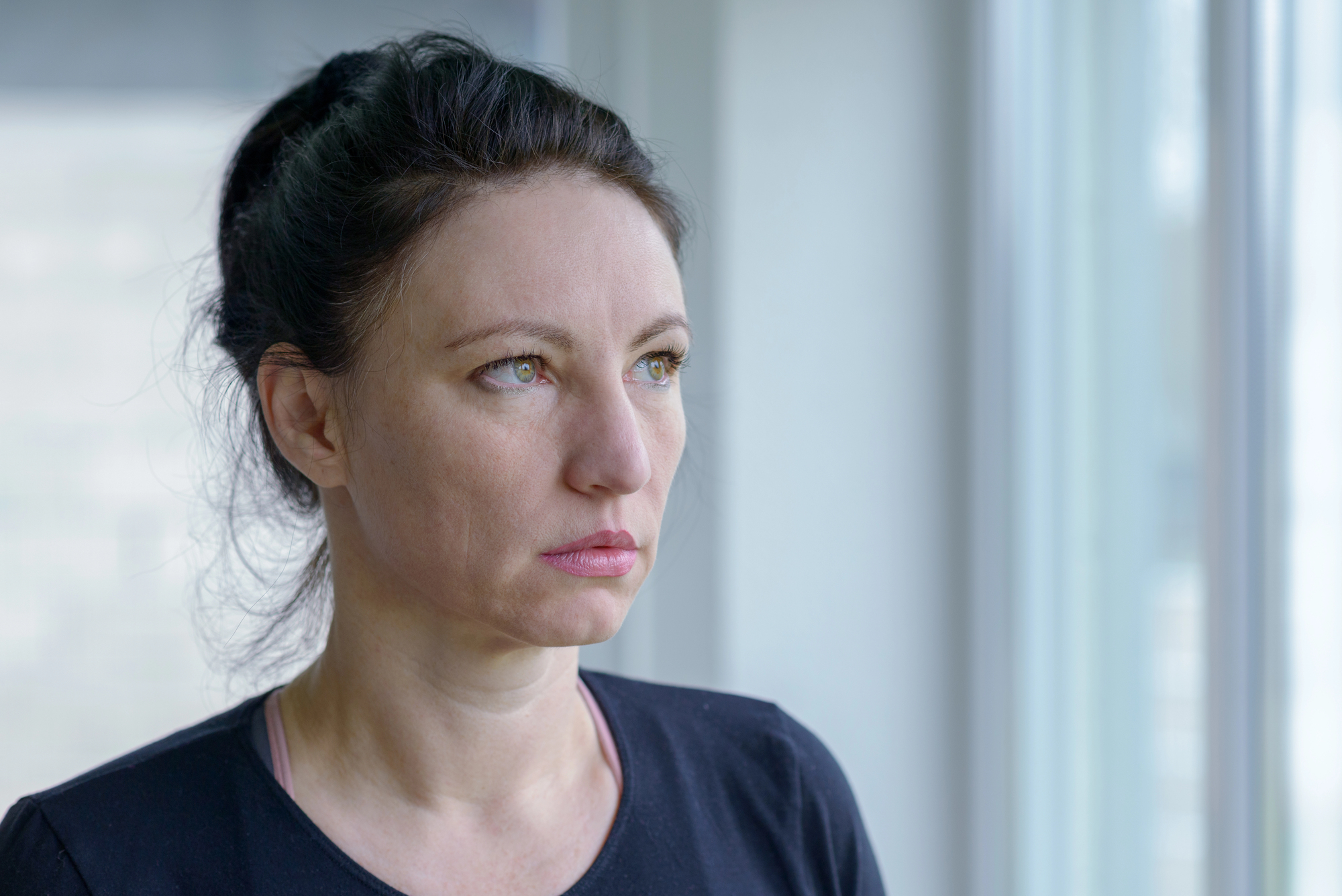 A woman with dark hair, tied back, stands by a window wearing a black top. She looks thoughtfully out the window, with soft natural light illuminating her face.