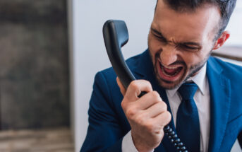 A man in a blue suit and white shirt is yelling into a black corded phone with an angry expression on his face. He is indoors with a blurred background, indicating an intense moment of frustration or anger during a phone call.