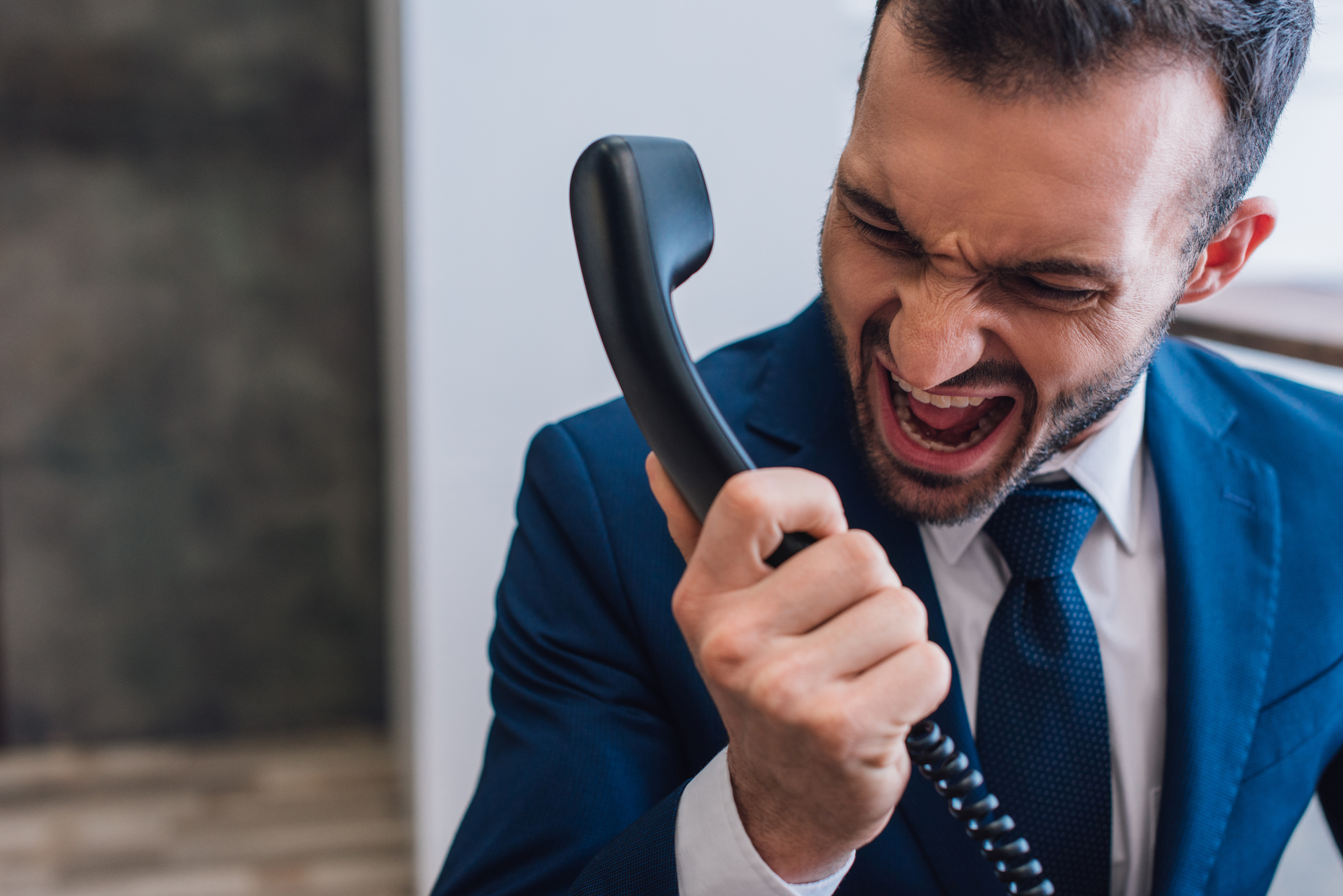 A man in a blue suit and white shirt is yelling into a black corded phone with an angry expression on his face. He is indoors with a blurred background, indicating an intense moment of frustration or anger during a phone call.