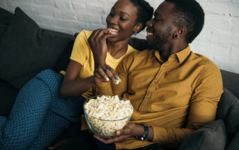 A couple sitting on a couch, smiling and sharing a bowl of popcorn. The woman is wearing a yellow shirt and blue patterned pants, while the man is wearing a mustard yellow shirt. They are enjoying a relaxed moment together in a cozy setting.