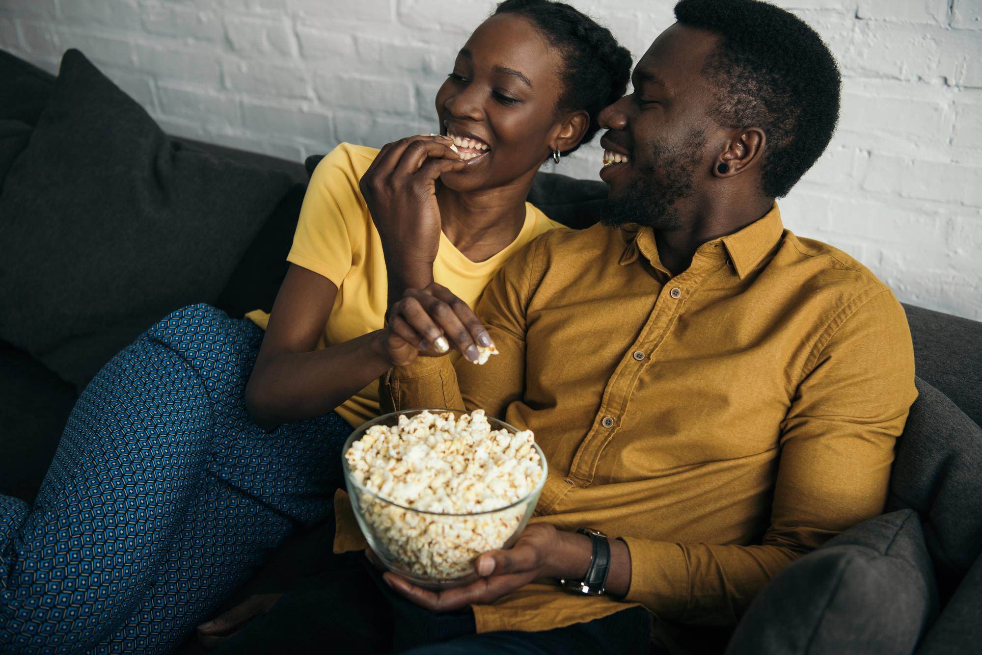 A couple sitting on a couch, smiling and sharing a bowl of popcorn. The woman is wearing a yellow shirt and blue patterned pants, while the man is wearing a mustard yellow shirt. They are enjoying a relaxed moment together in a cozy setting.