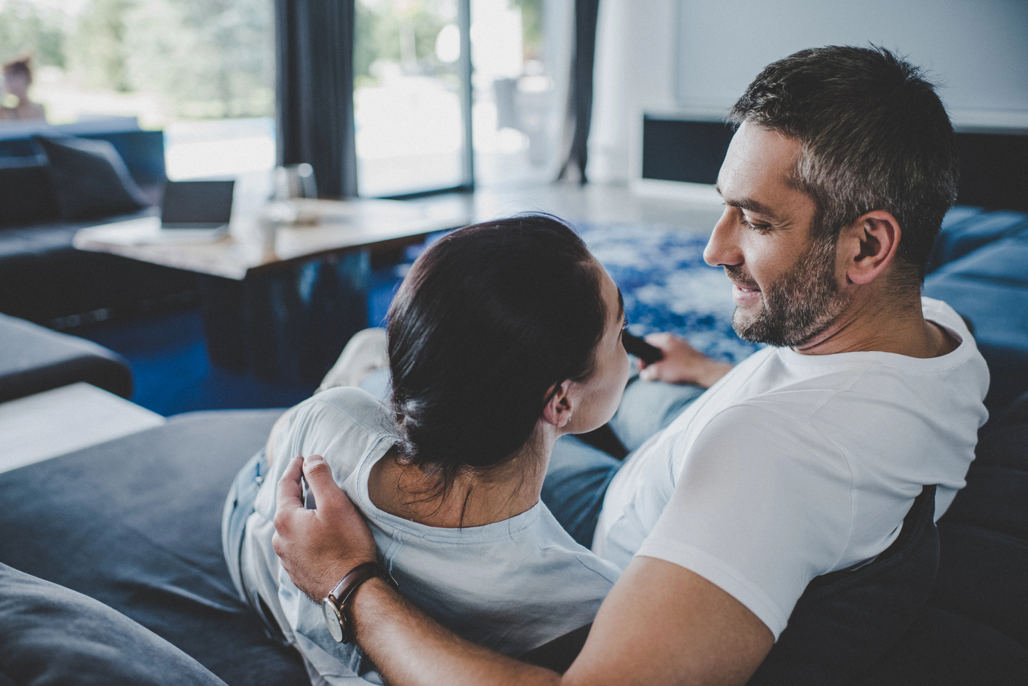 A couple sits closely on a couch in a cozy living room, with soft natural light coming through large windows in the background. They are comfortably embracing and smiling at each other. A laptop is open on a table nearby.