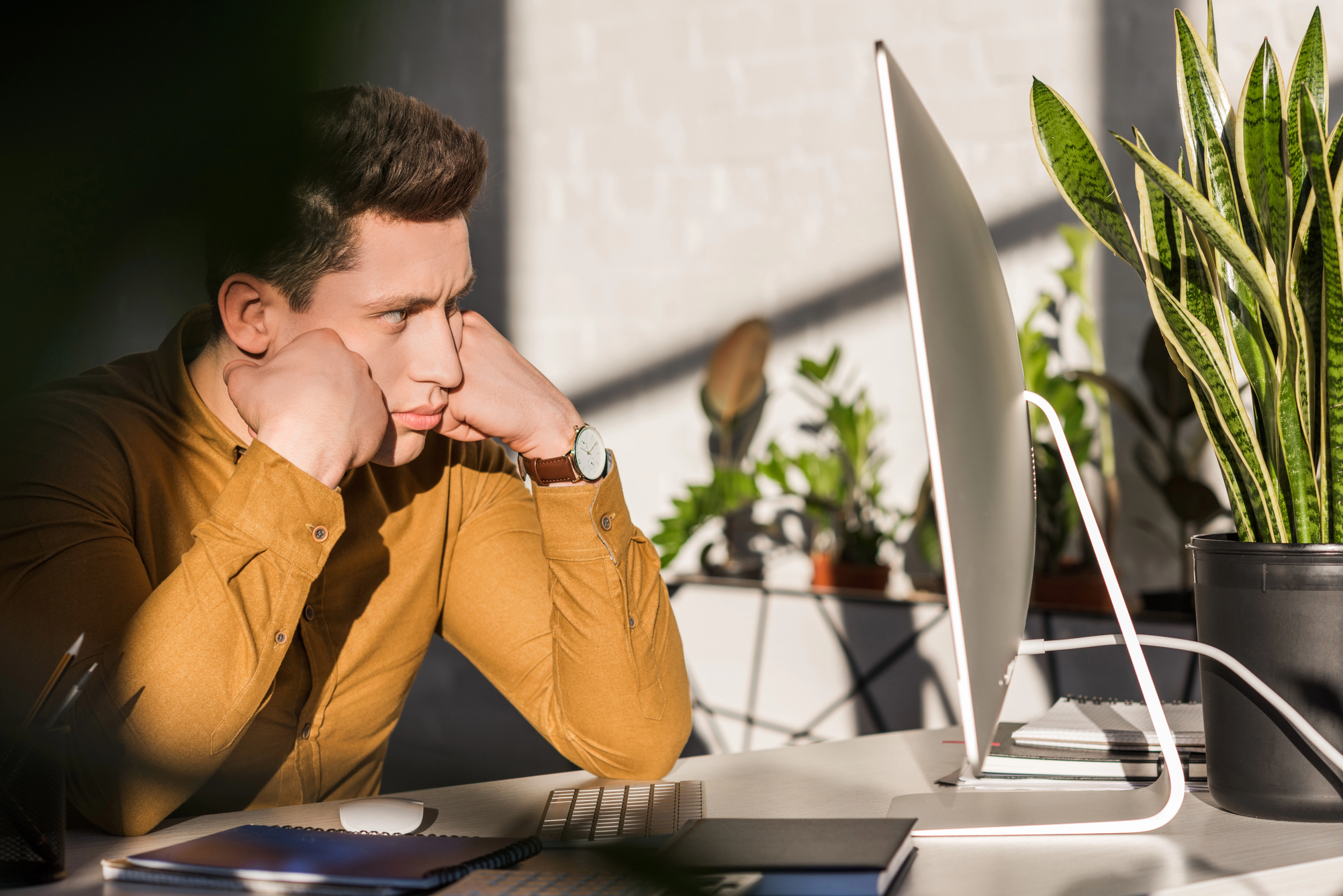 A person in a mustard-colored shirt sits at a desk, resting their face on their hands, looking thoughtfully at a computer monitor. Sunlight and plants are in the background, creating a tranquil office environment.